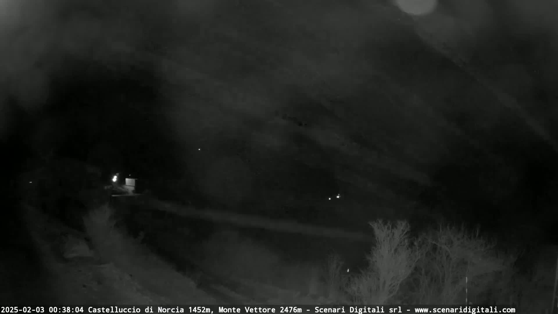 A nighttime, low-light view of a dark, hilly landscape with sparse vegetation under a cloudy sky.