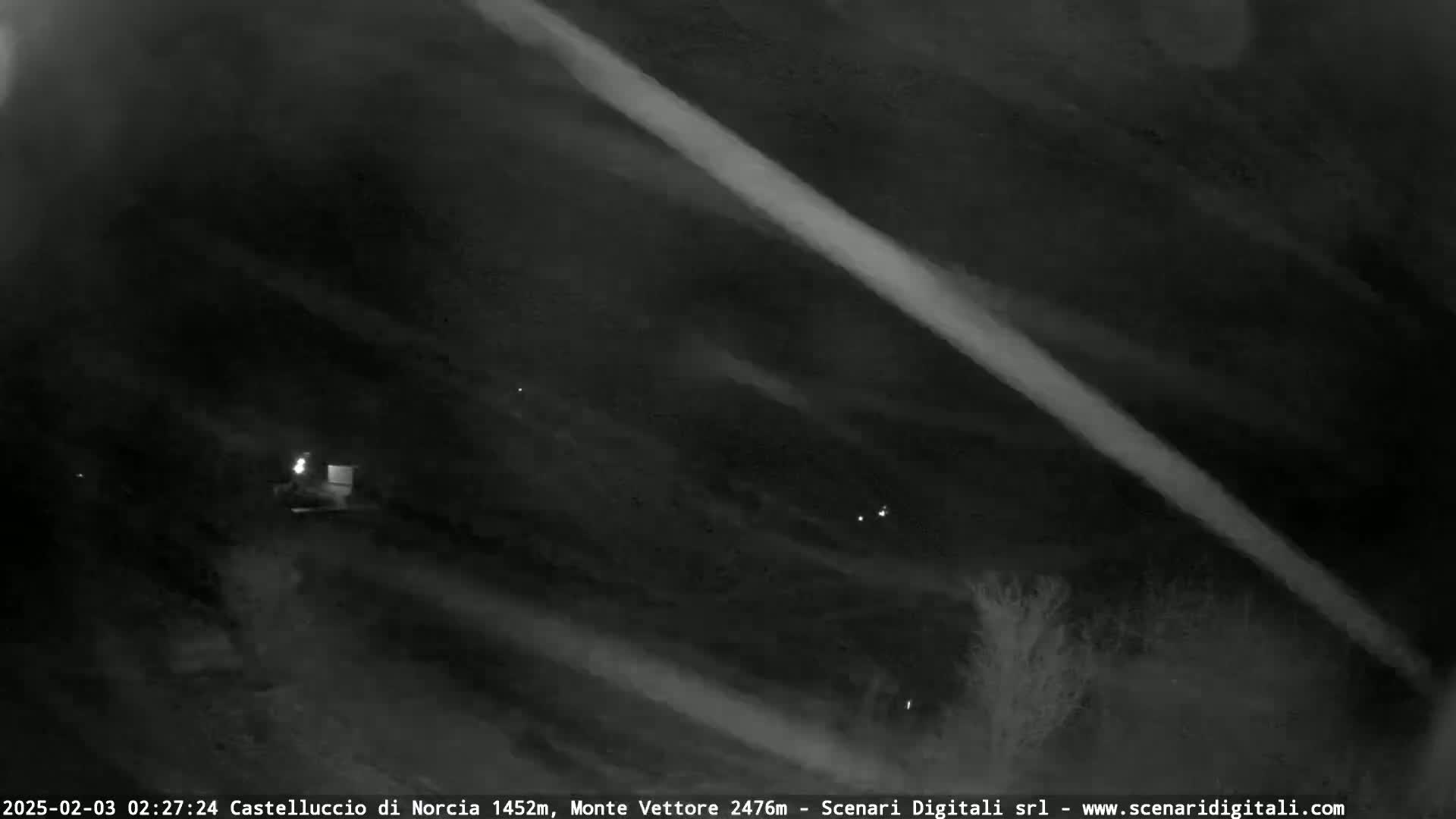 A nighttime, low-light image shows a dark landscape with faint streaks of light across the sky and a few small points of light possibly from distant buildings or lights.
