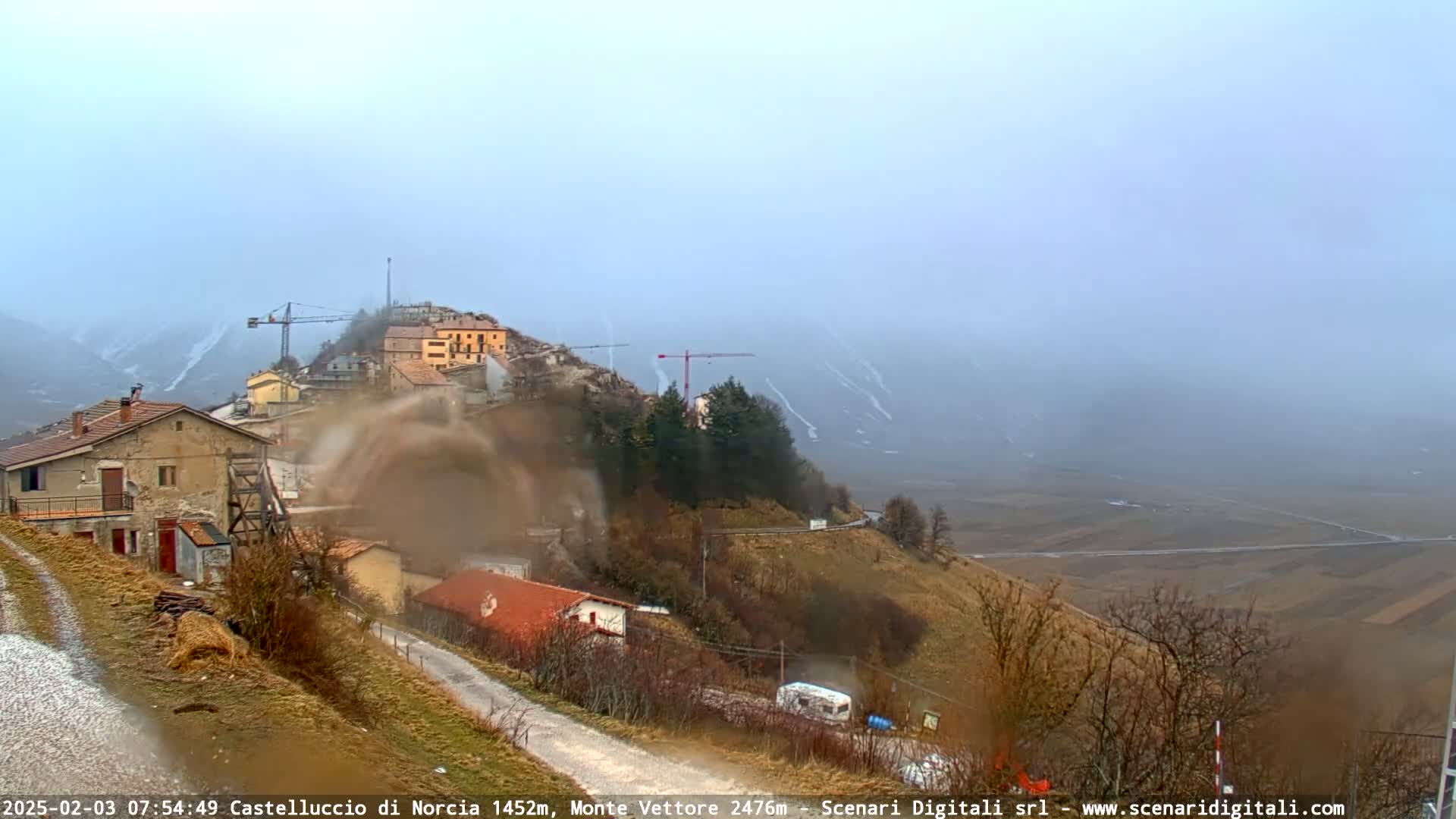 A hazy, possibly rainy, view of a small village nestled on a hillside, with construction cranes visible and a valley stretching out into the distance.