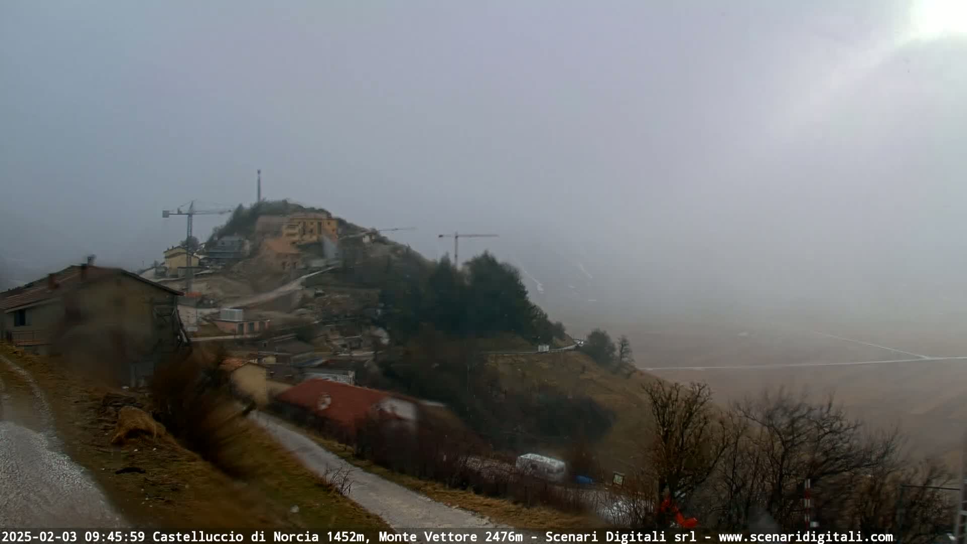 A foggy, rainy view of a hillside village with construction cranes visible on a hilltop in the distance.