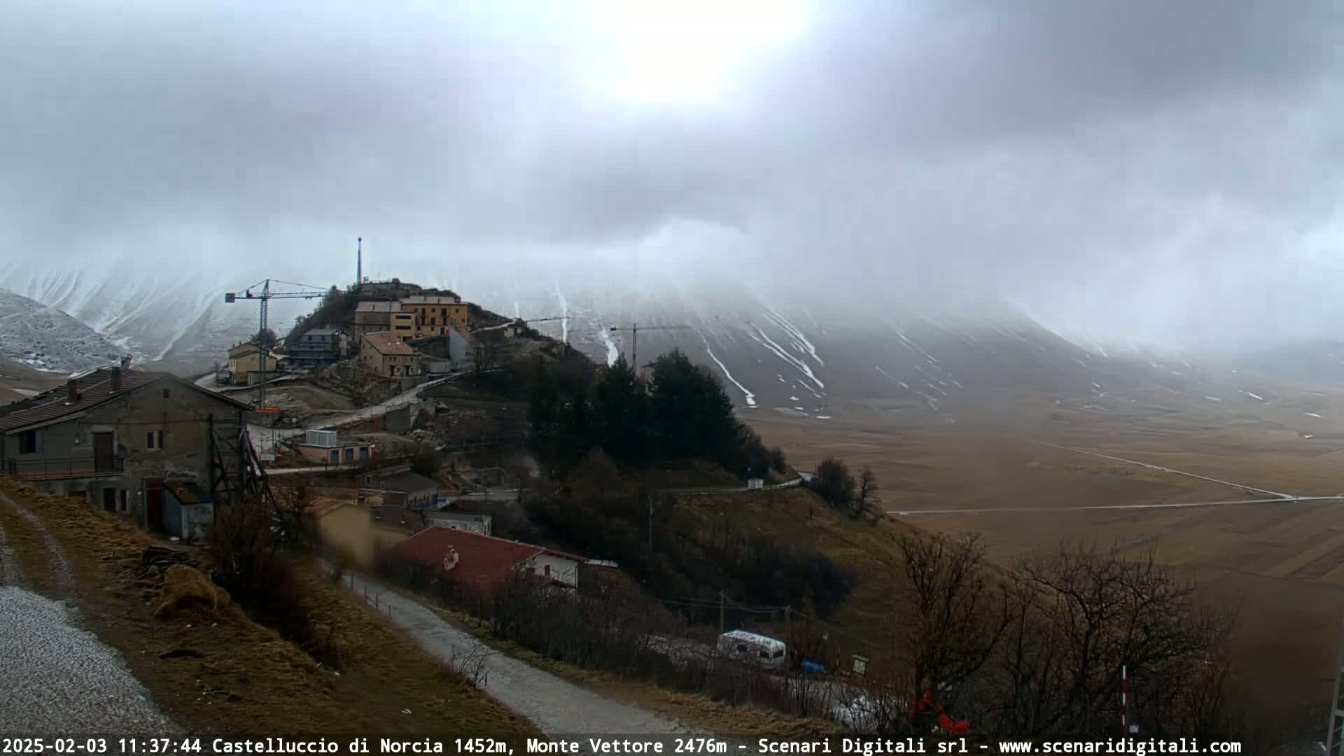 A small village nestled on a hillside, with construction cranes visible, overlooks a snow-dusted valley under a cloudy, snowy sky.