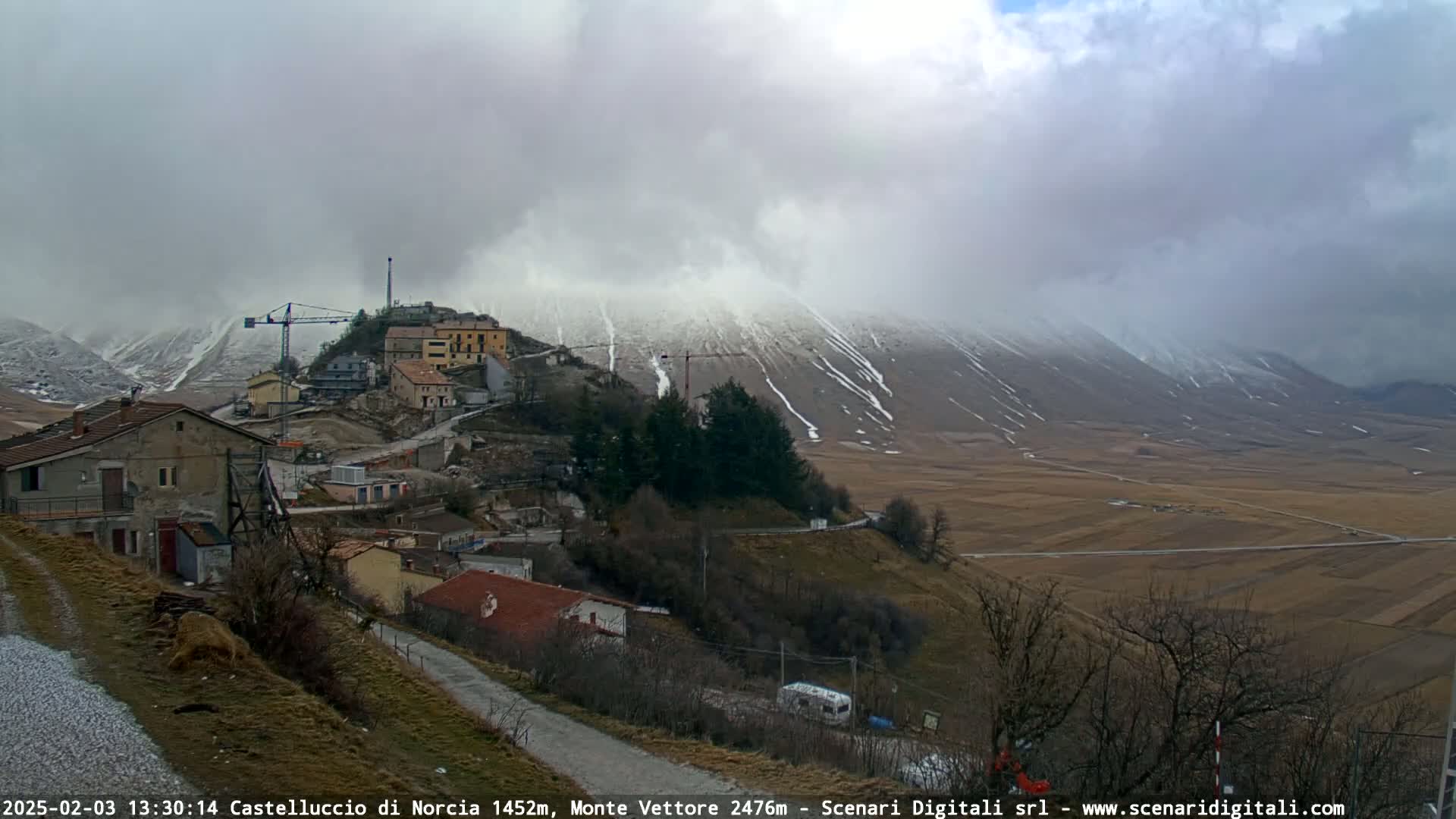 A small village with a construction crane is nestled in a valley below a snow-dusted mountain range under a cloudy sky.
