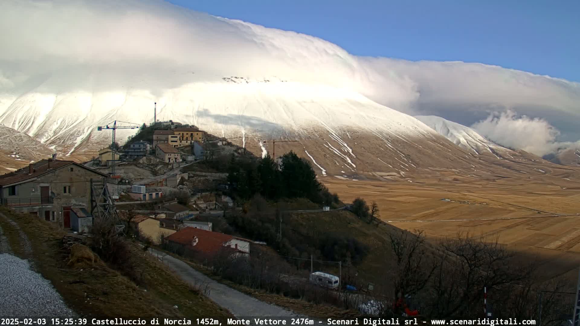 A small village nestled on a hillside overlooks a snow-dusted mountain under a partly cloudy sky.