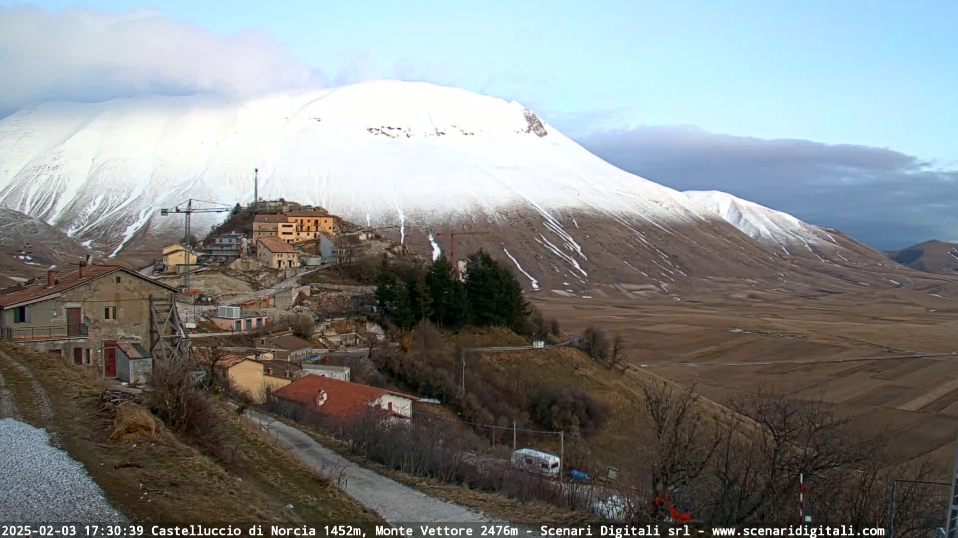 A snow-covered mountain looms over a small village nestled in a valley, under a partly cloudy sky.