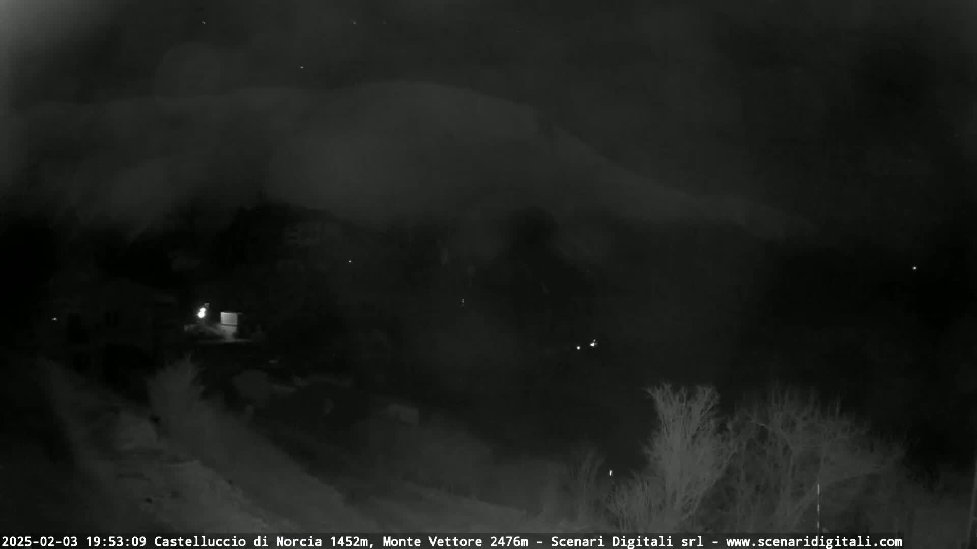A nighttime, low-light view of a dark, mountainous landscape with sparse vegetation and a few faint lights visible in the distance.