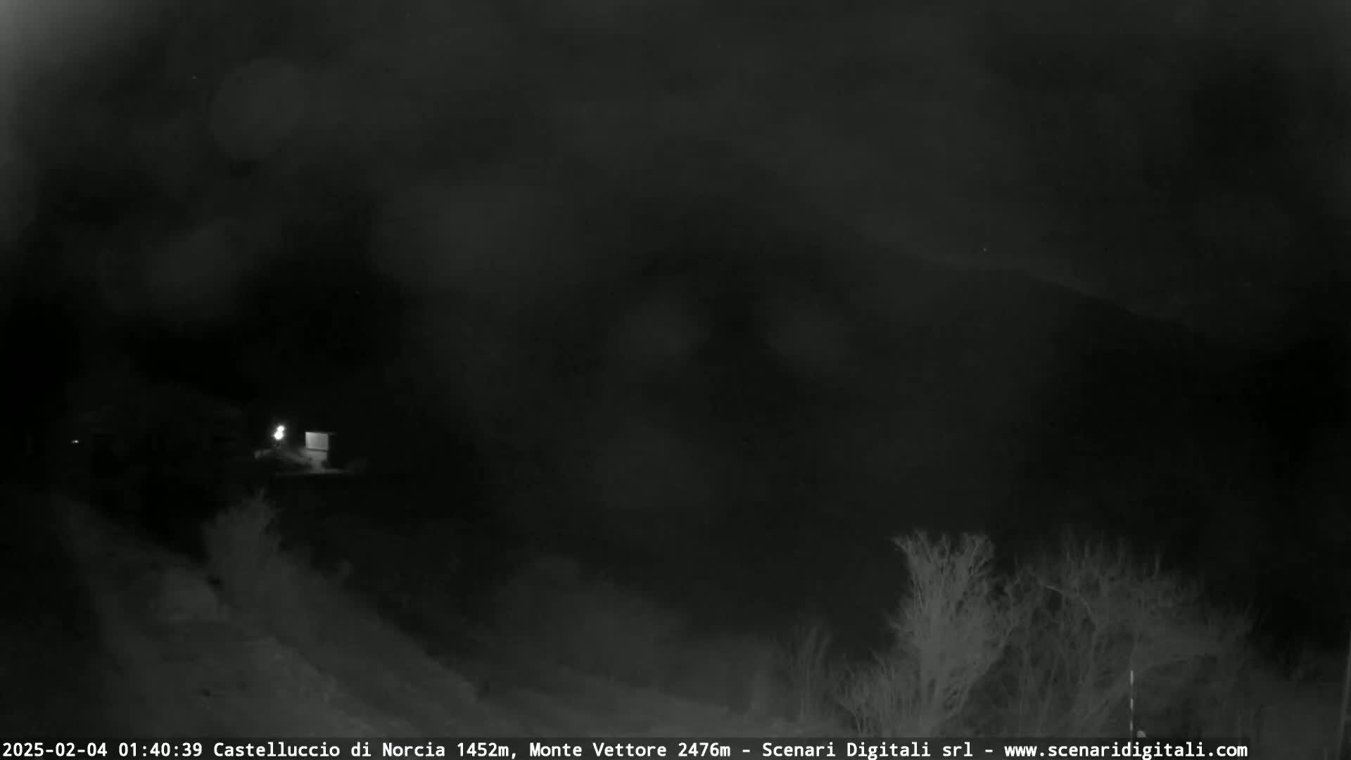 A nighttime, low-light view of a dark landscape with sparse, silhouetted trees and a dimly lit structure in the mid-ground, under a cloudy sky.