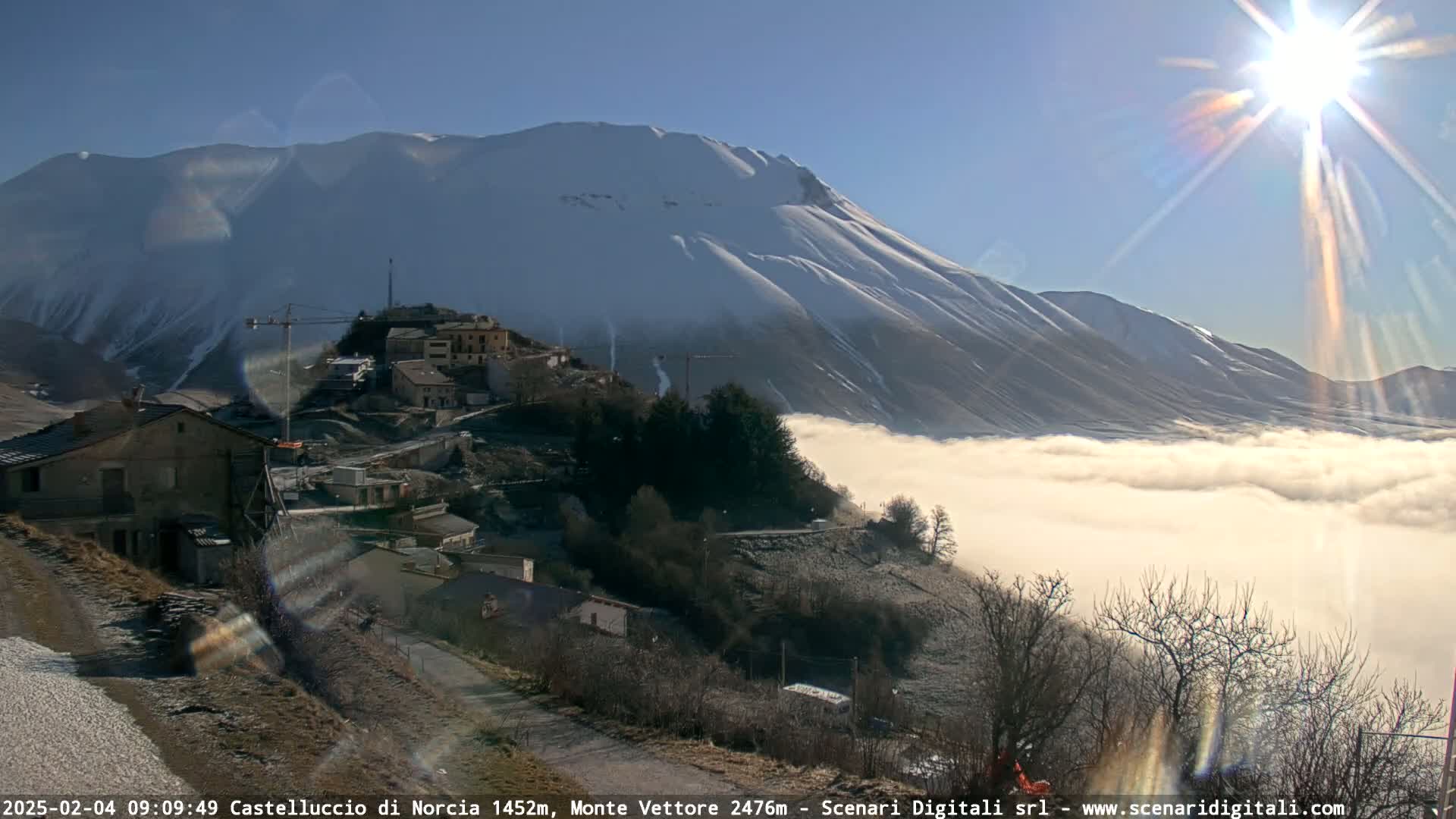 A snow-covered mountain overlooks a small village nestled in a valley filled with fog, under a bright, sunny sky.