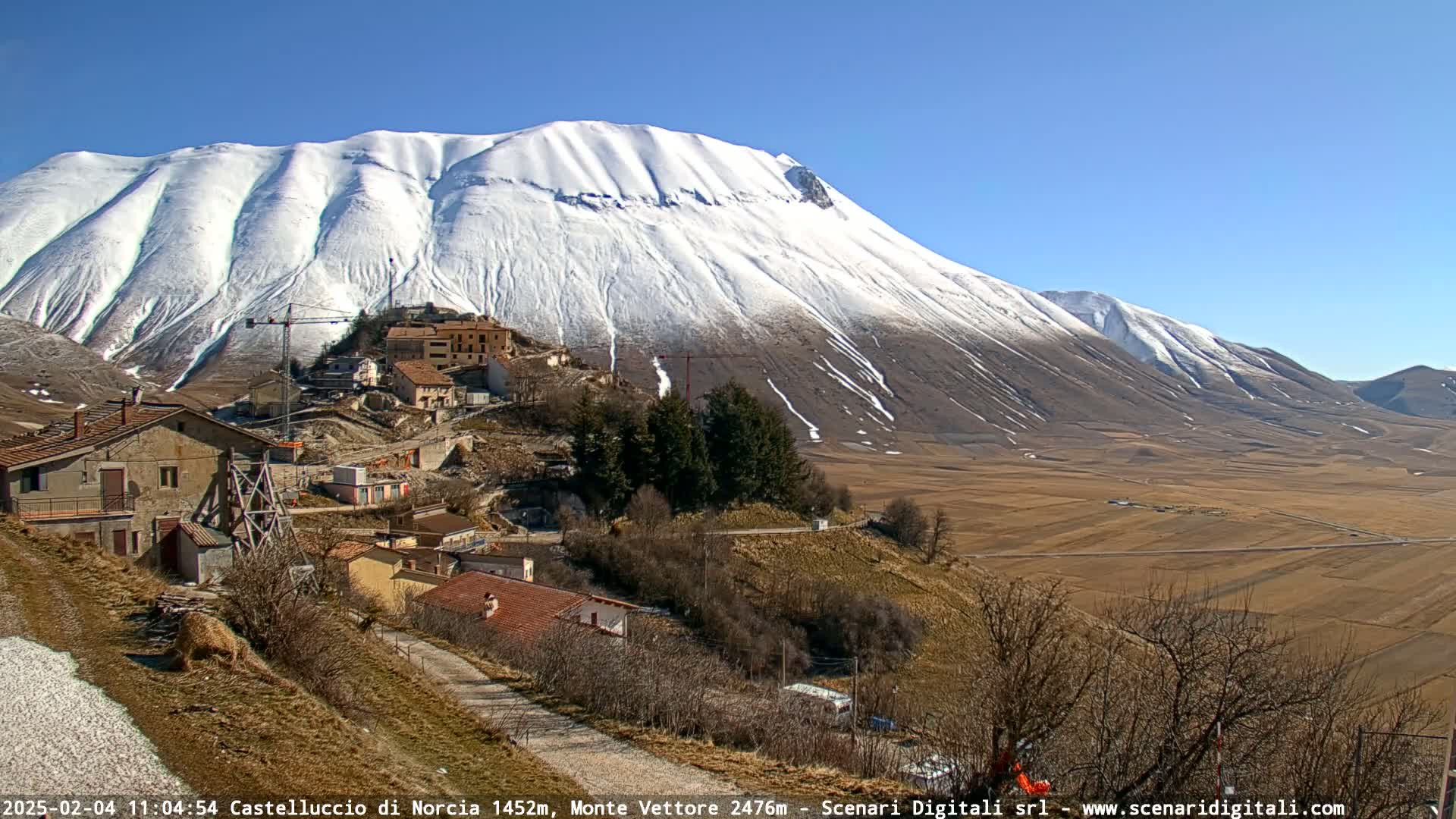 A snow-covered mountain looms over a small village nestled in a valley on a sunny day.