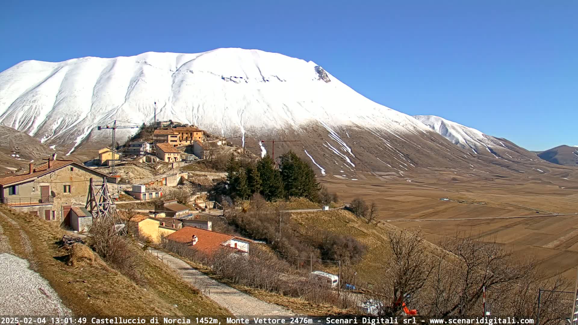 A snow-capped mountain overlooks a small village nestled in a valley under a clear blue sky.