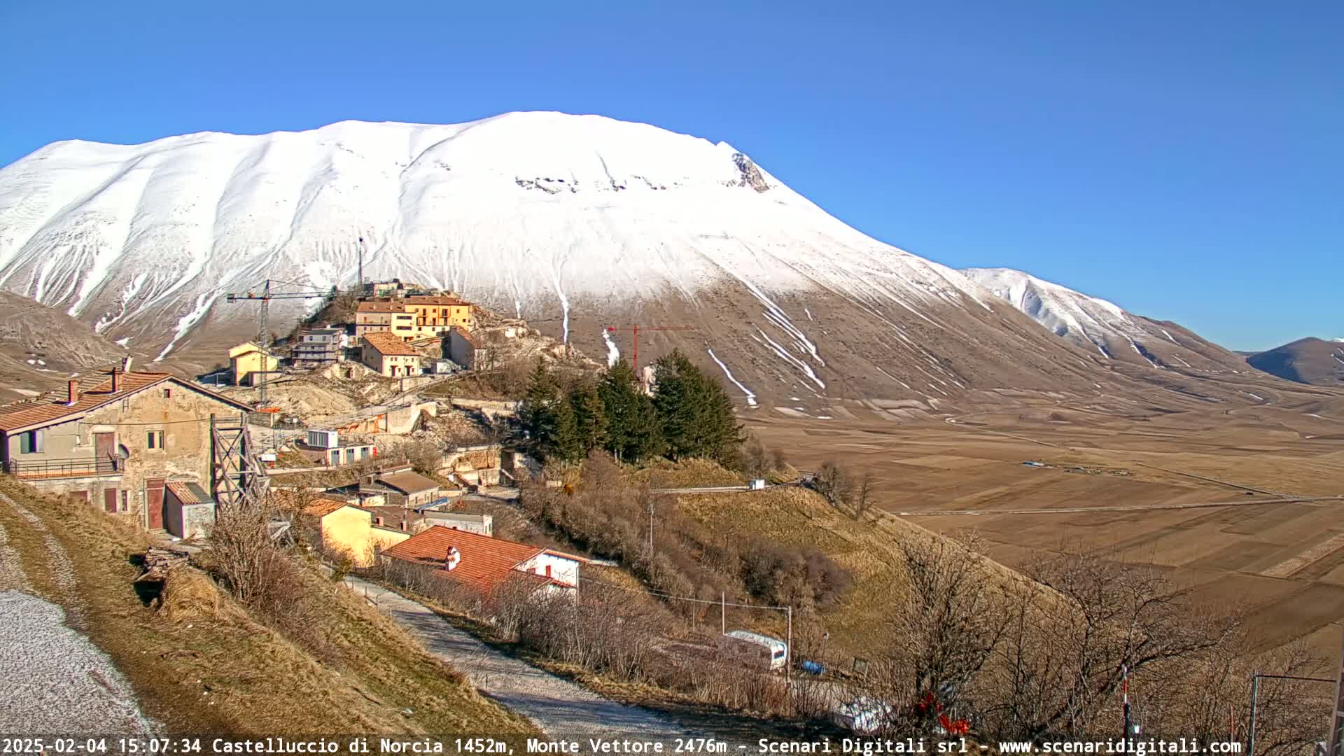 A snow-capped mountain overlooks a small village nestled in a valley under a clear blue sky.
