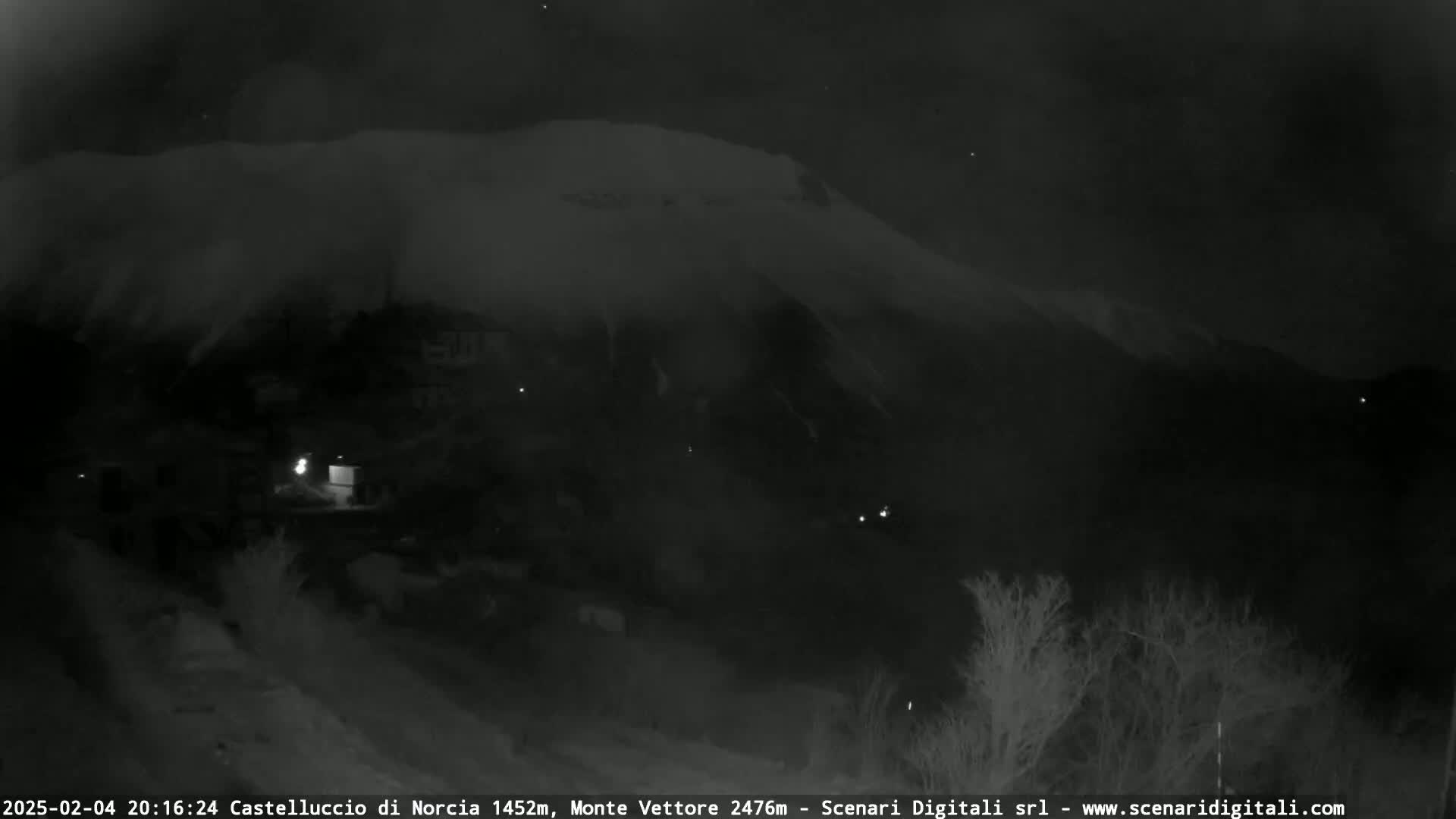 A nighttime, snowy mountain overlooks a small, dimly lit town under a dark sky.