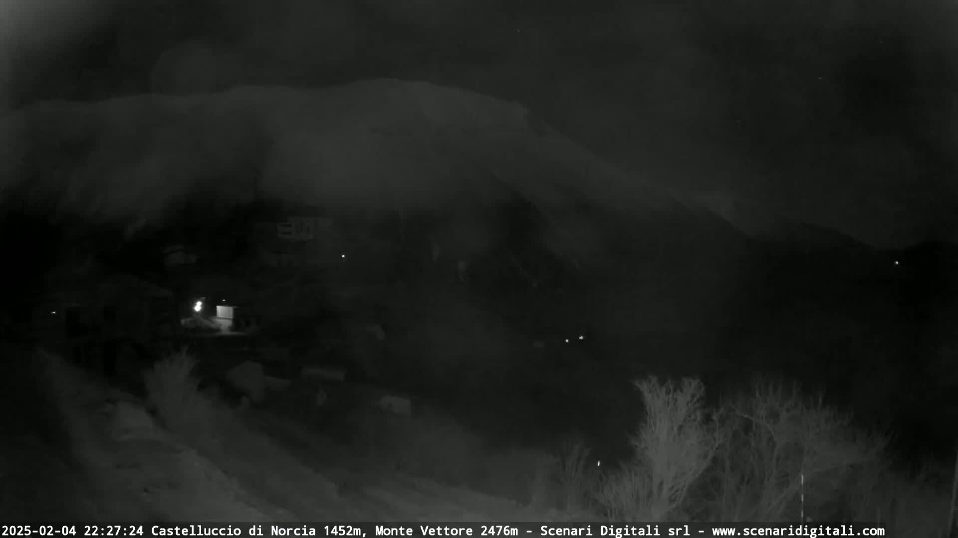 A nighttime, low-light view of a snow-covered mountain and a sparsely lit town below, under a cloudy sky.