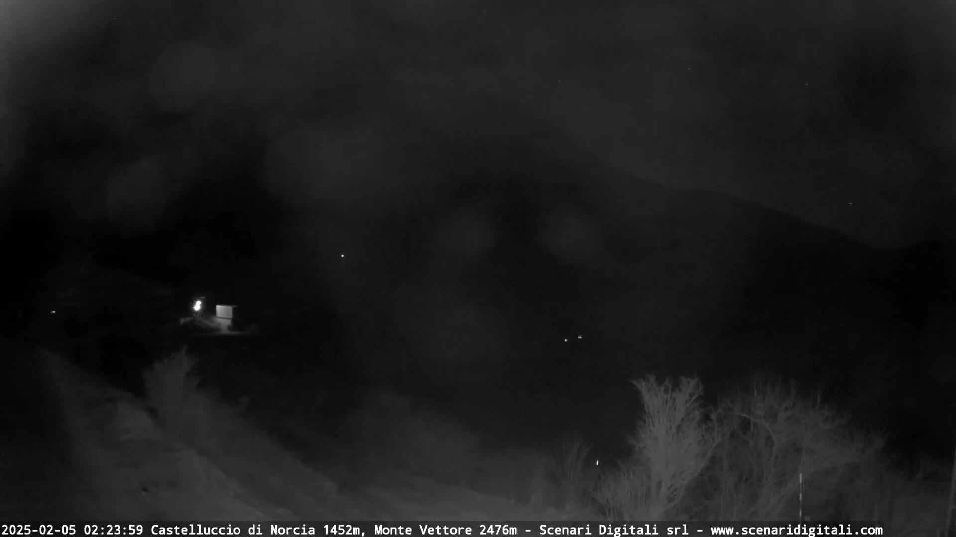A nighttime, low-light view shows a dark landscape with a few distant lights and some silhouetted trees in the foreground under a mostly cloudy sky.