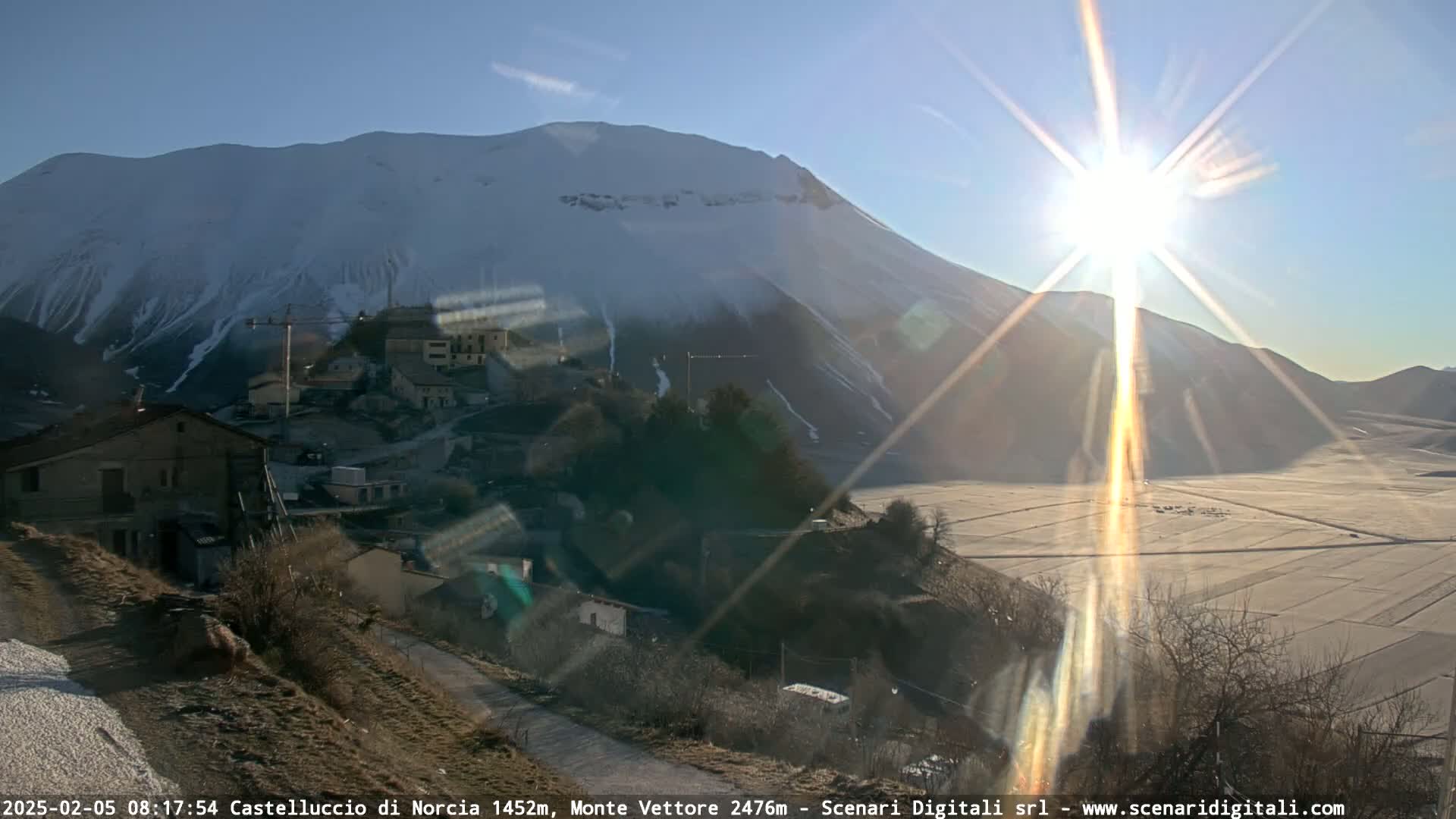 A sunlit village nestled below a snow-capped mountain on a clear, bright day.