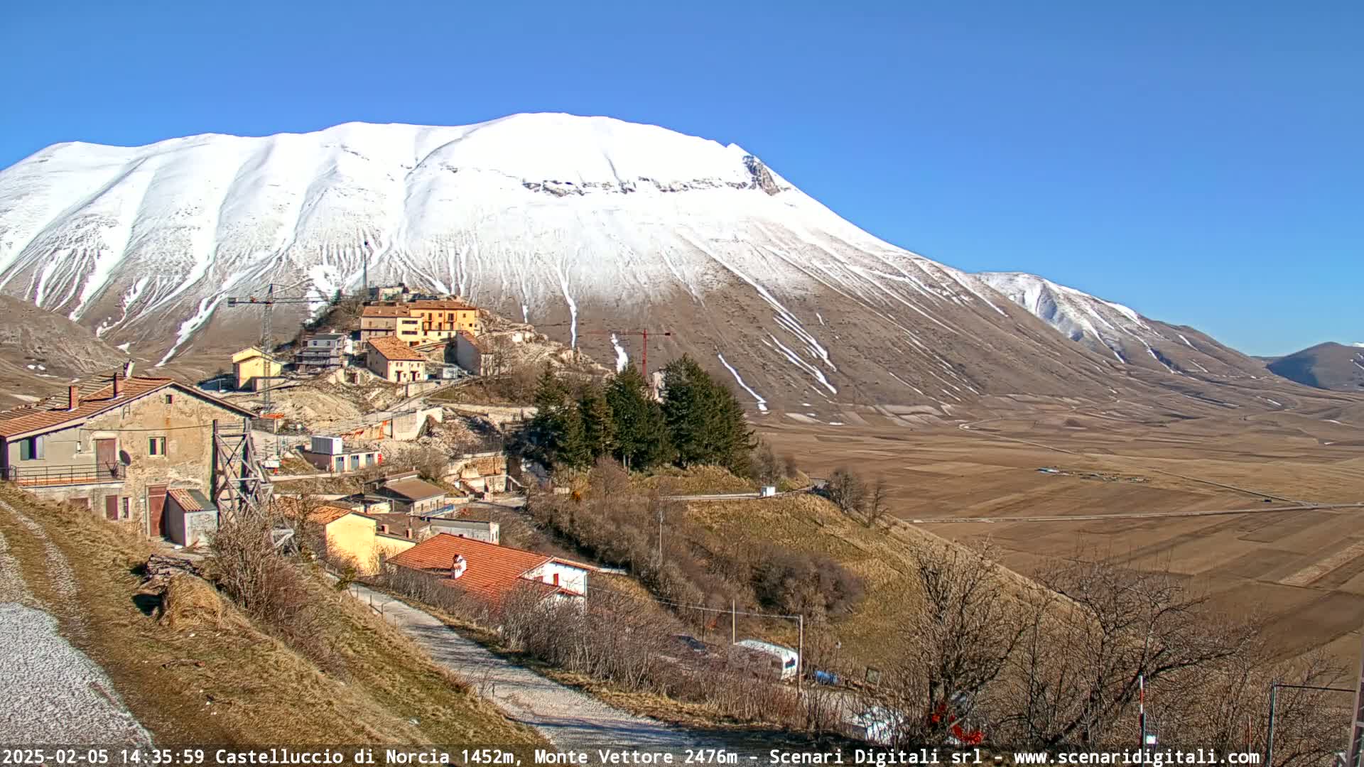 A snow-capped mountain overlooks a small village nestled in a valley on a sunny day.
