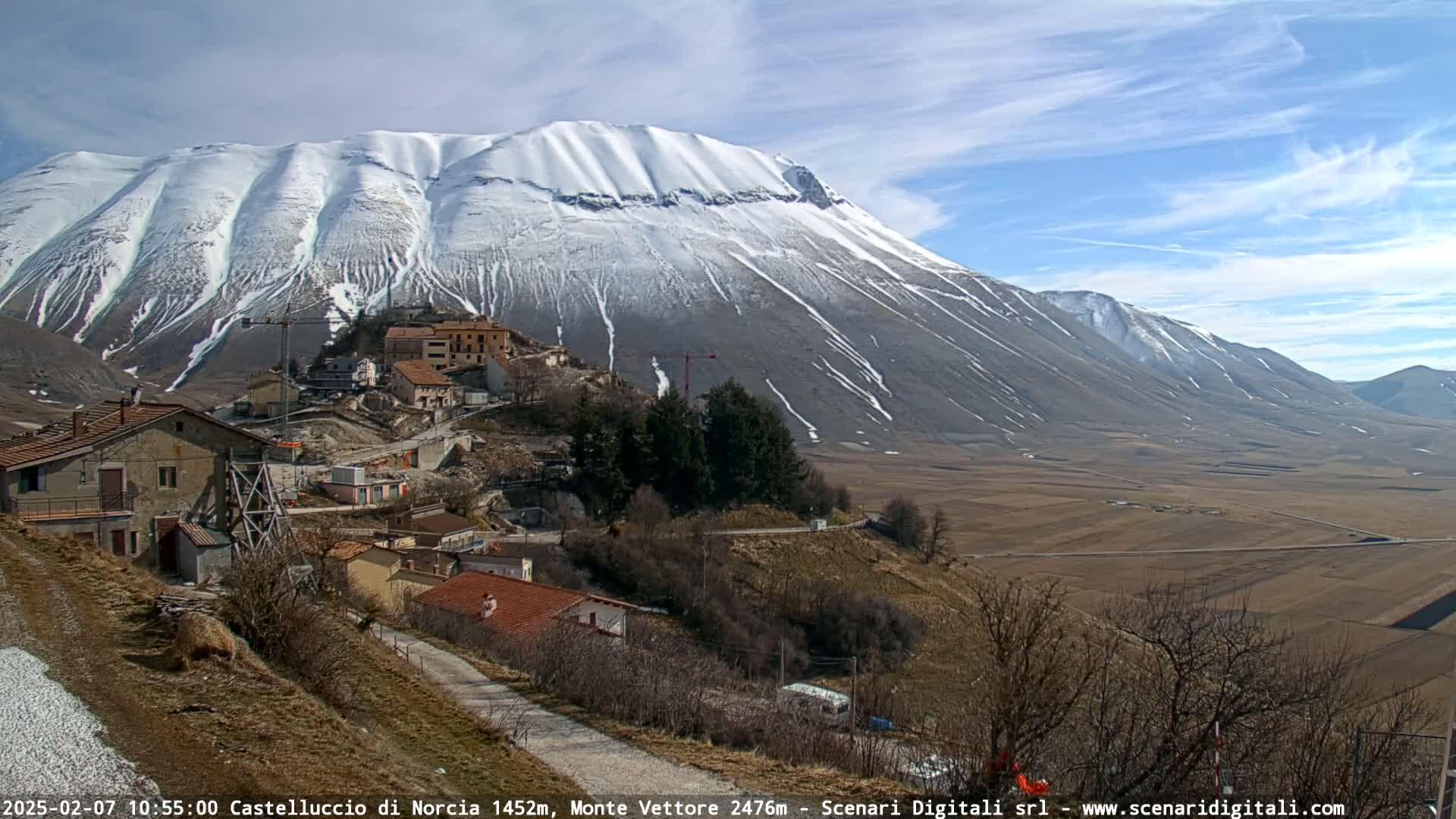 A snow-capped mountain overlooks a small village nestled in a valley under a mostly clear, sunny sky.