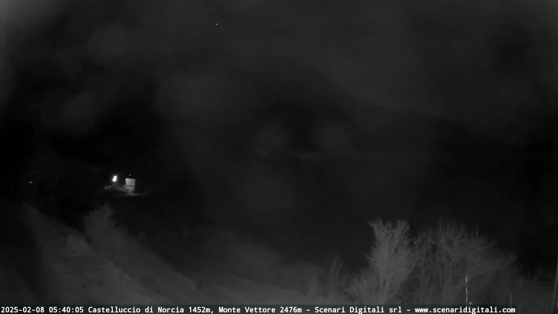 A nighttime, dark and cloudy scene shows a small building and some vegetation.