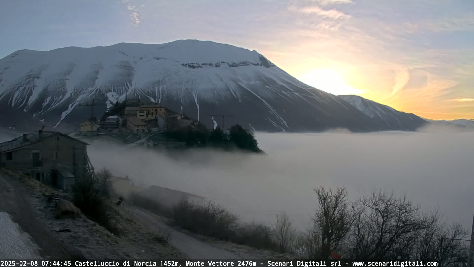 A snow-capped mountain at sunrise is partially obscured by a thick layer of fog covering a valley below, where a small village sits nestled among bare trees.