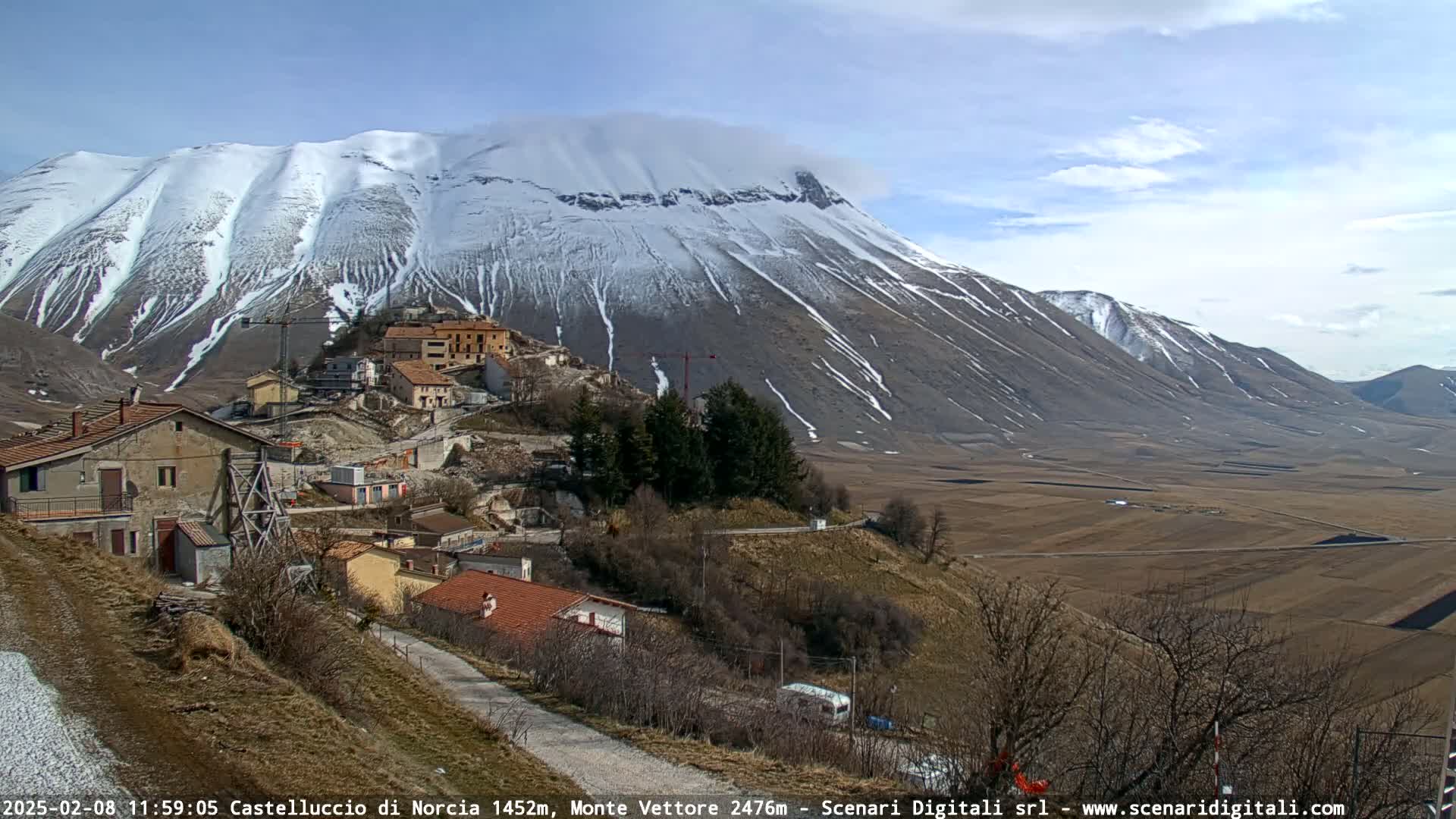 A snow-capped mountain overlooks a small village nestled in a valley of brown fields under a mostly clear sky.