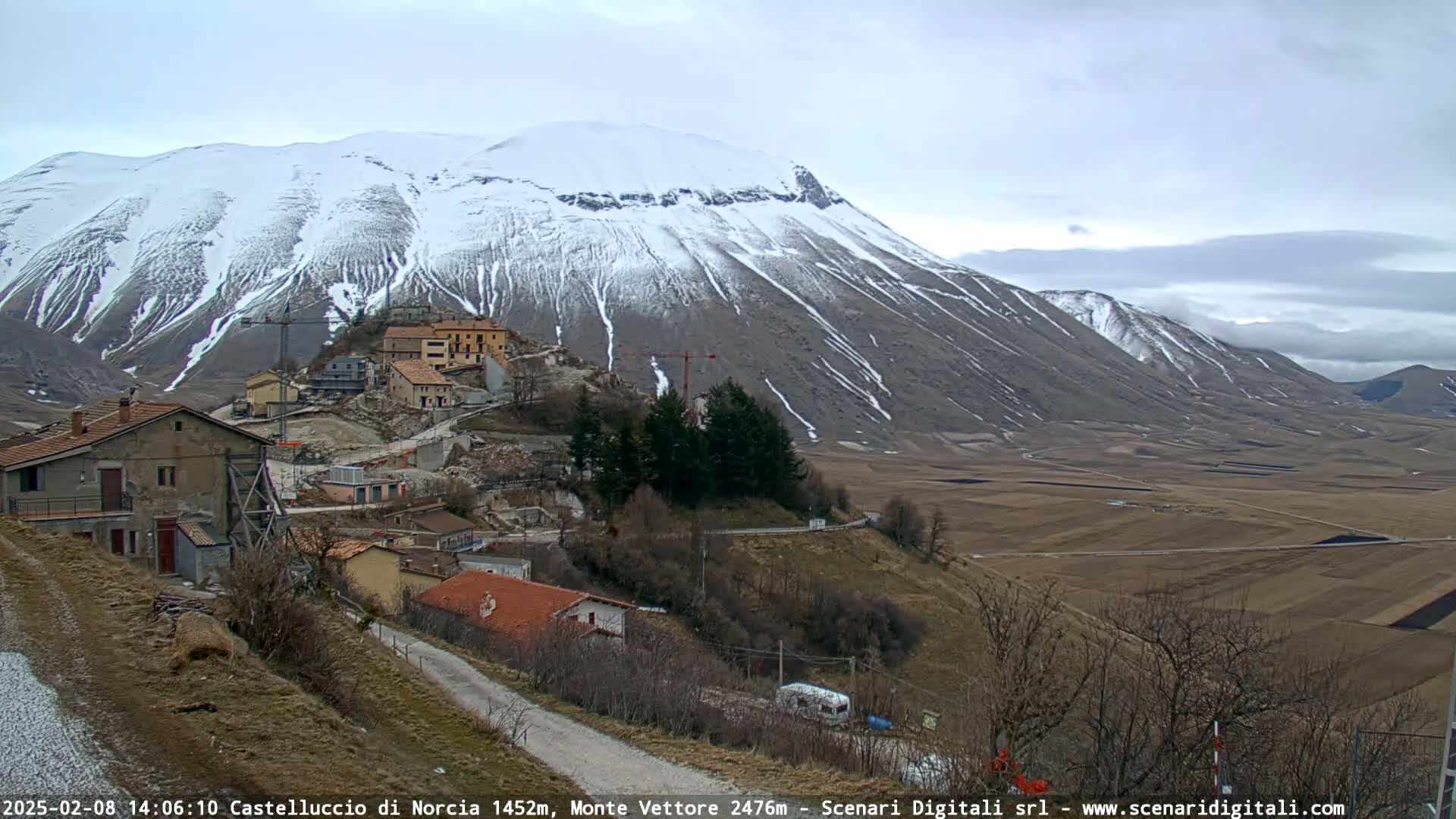A snow-dusted mountain overlooks a small village nestled in a valley under an overcast sky.