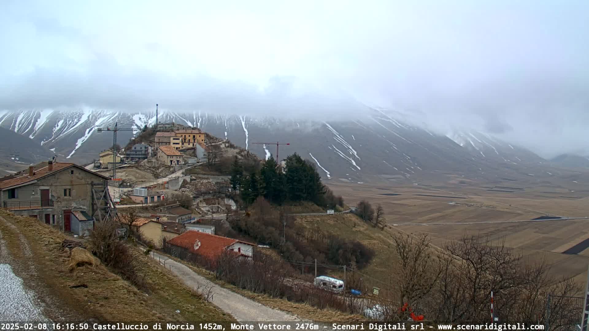 A hillside village with construction cranes is nestled below snow-dusted mountains under a cloudy sky.