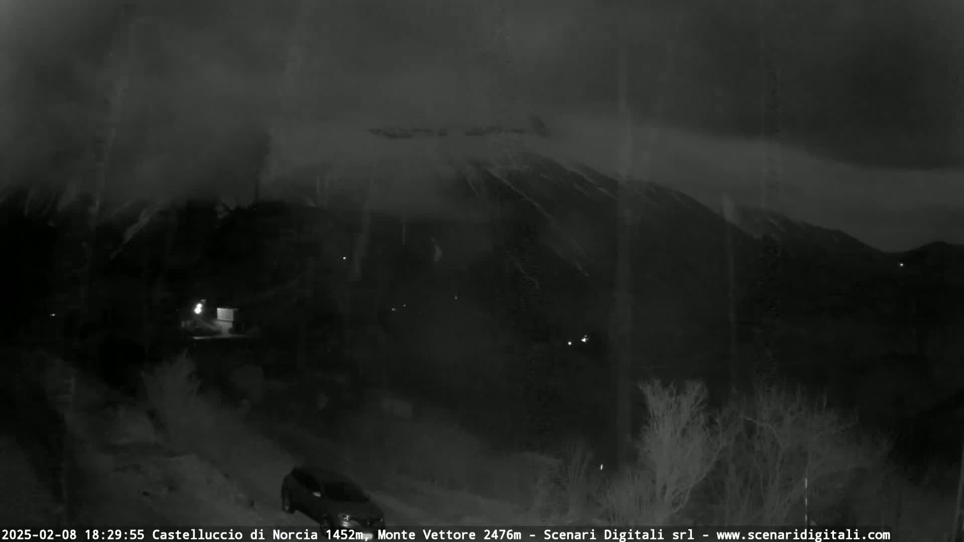 A nighttime, overcast view shows a dark mountain range under a cloudy sky, with a few faint lights visible in the foreground.