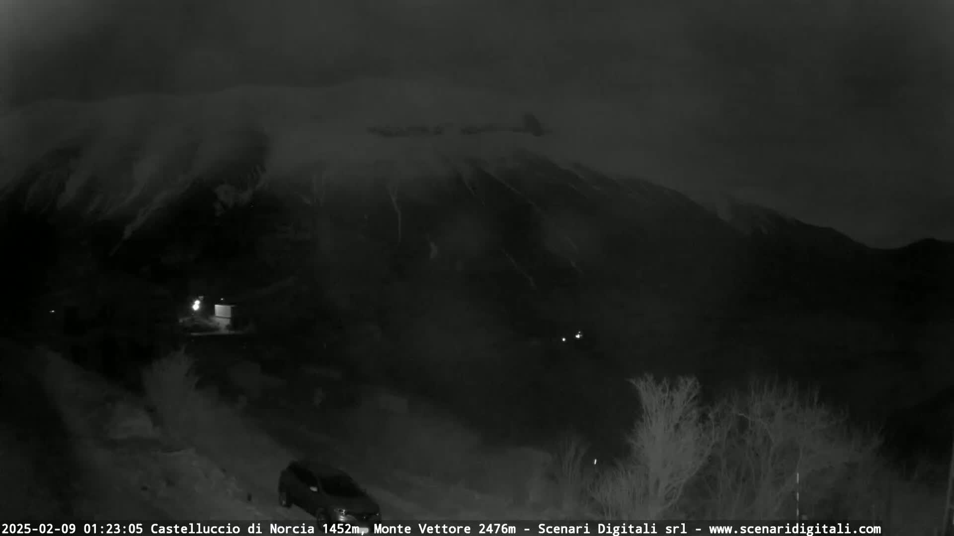 A nighttime, snowy scene shows a mountain range under a dark, cloudy sky, with sparsely vegetated land in the foreground.