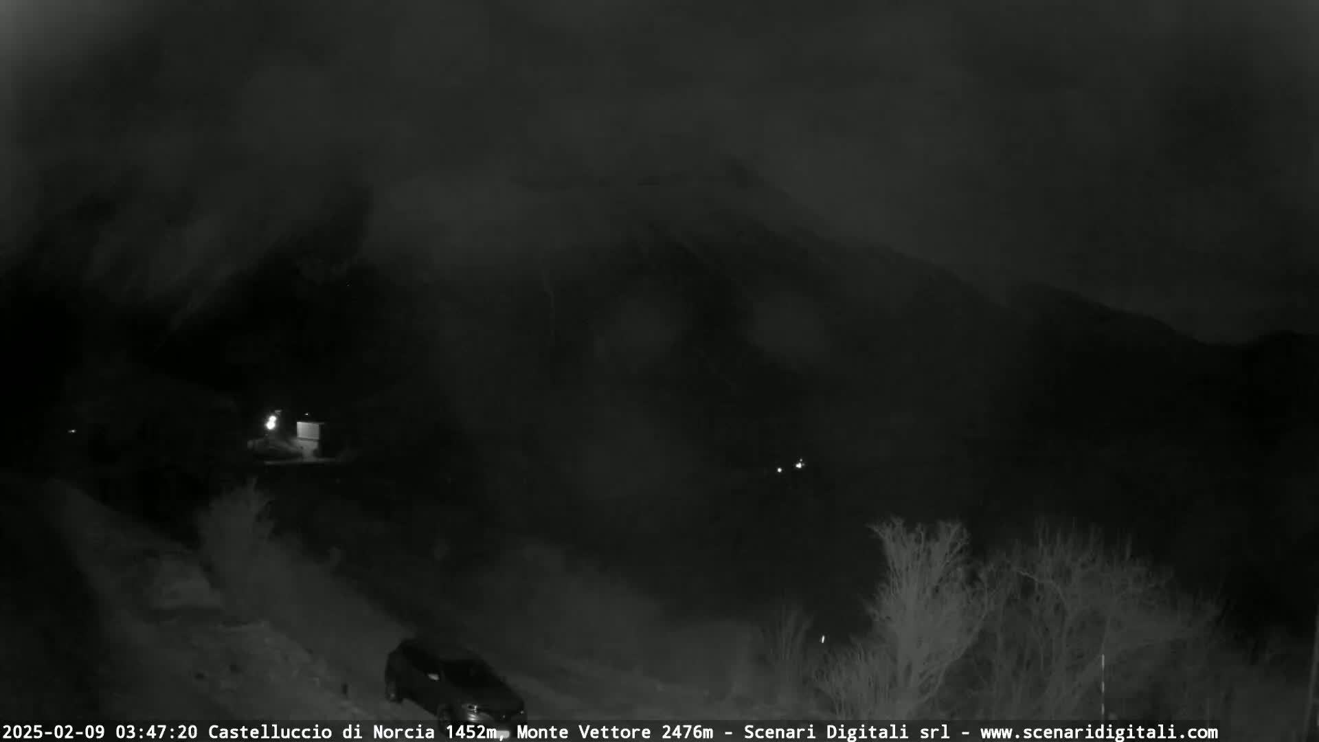 A nighttime, dark, and hazy view of a mountain range under a cloudy sky, with a few faint lights visible in the distance.