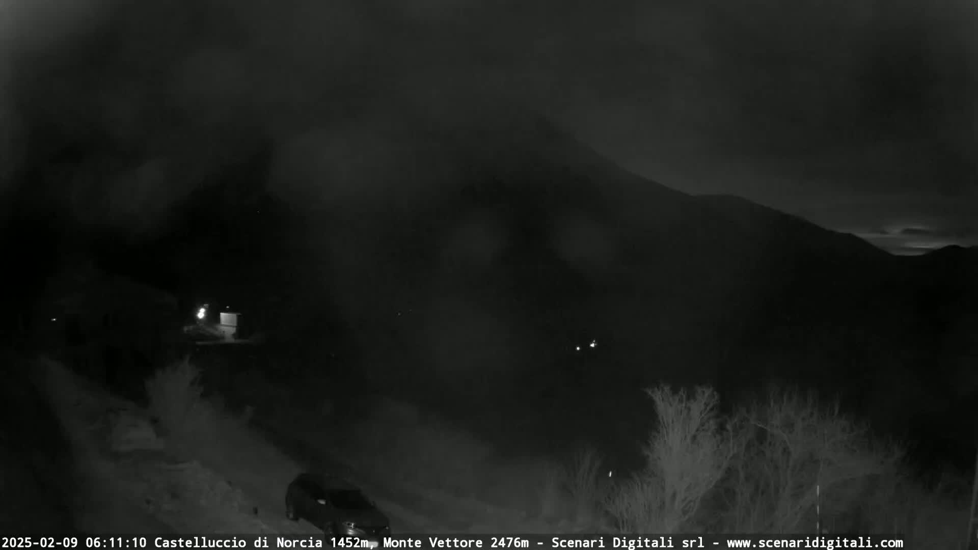 A nighttime, snowy scene shows a dark mountain range under a cloudy sky, with a few lights visible in the foreground and a vehicle parked near the bottom.