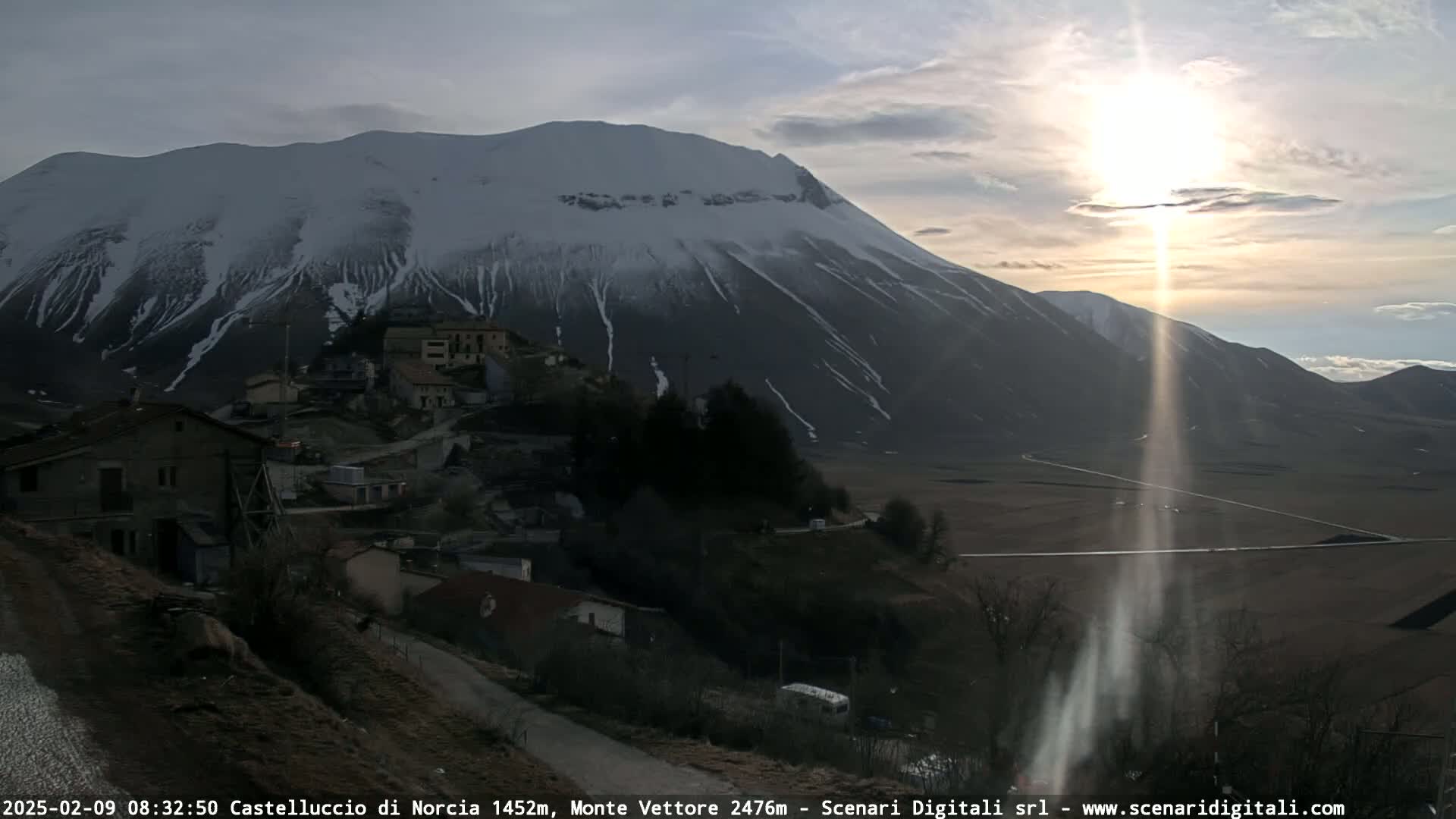 A sunlit valley features a snow-capped mountain in the background, overlooking a small village nestled in the foothills.