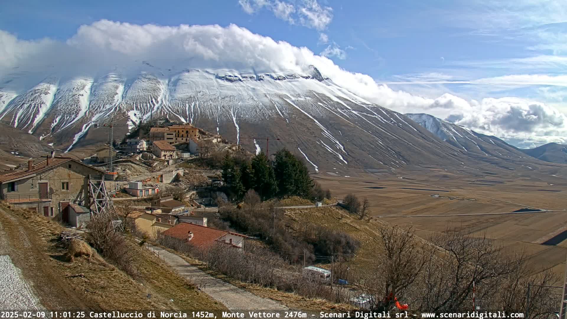 A snow-capped mountain range stretches across a sunny sky above a small village nestled in a valley of brown farmland.