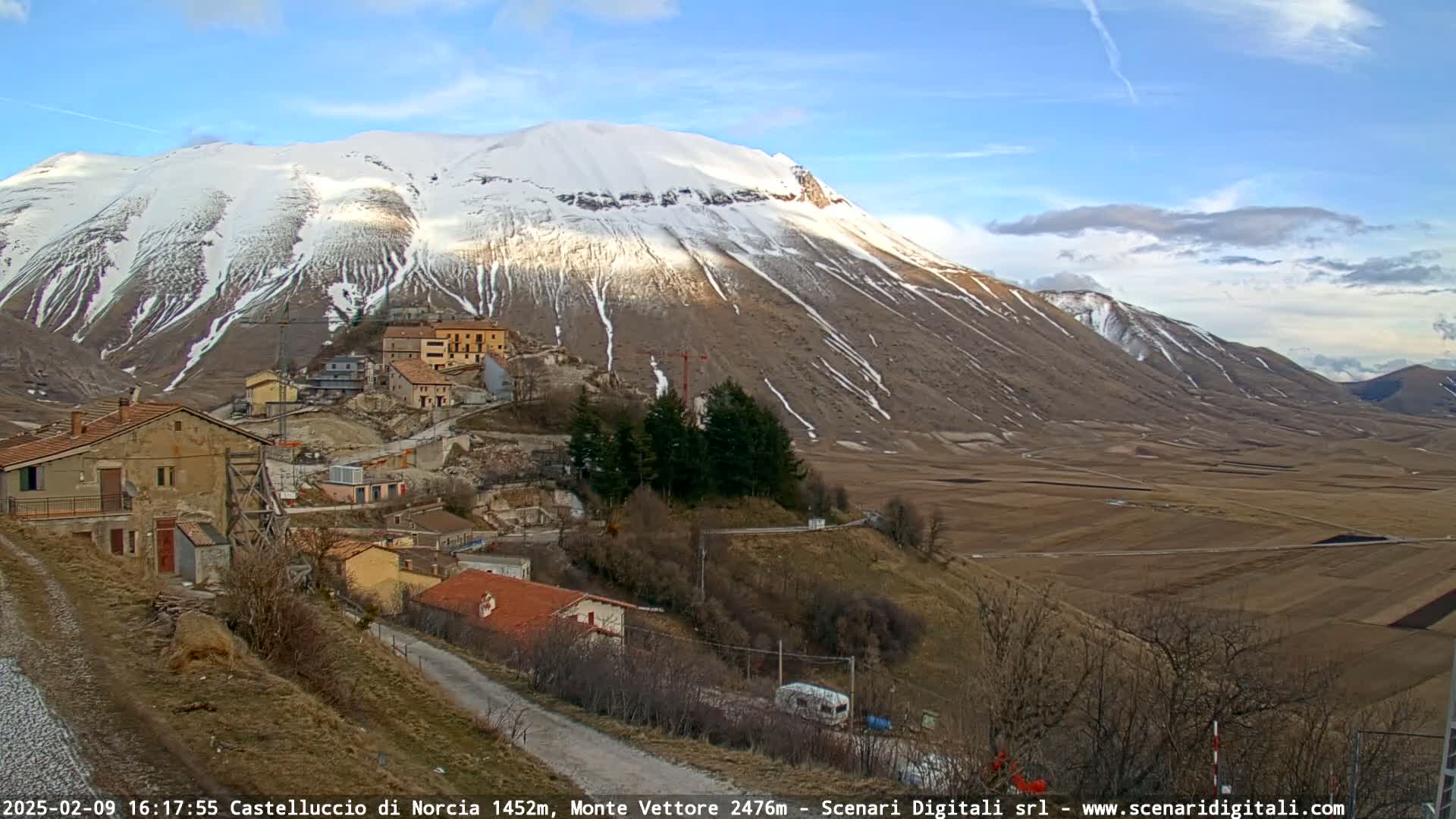 A snow-capped mountain overlooks a small village nestled in a valley on a partly cloudy day.