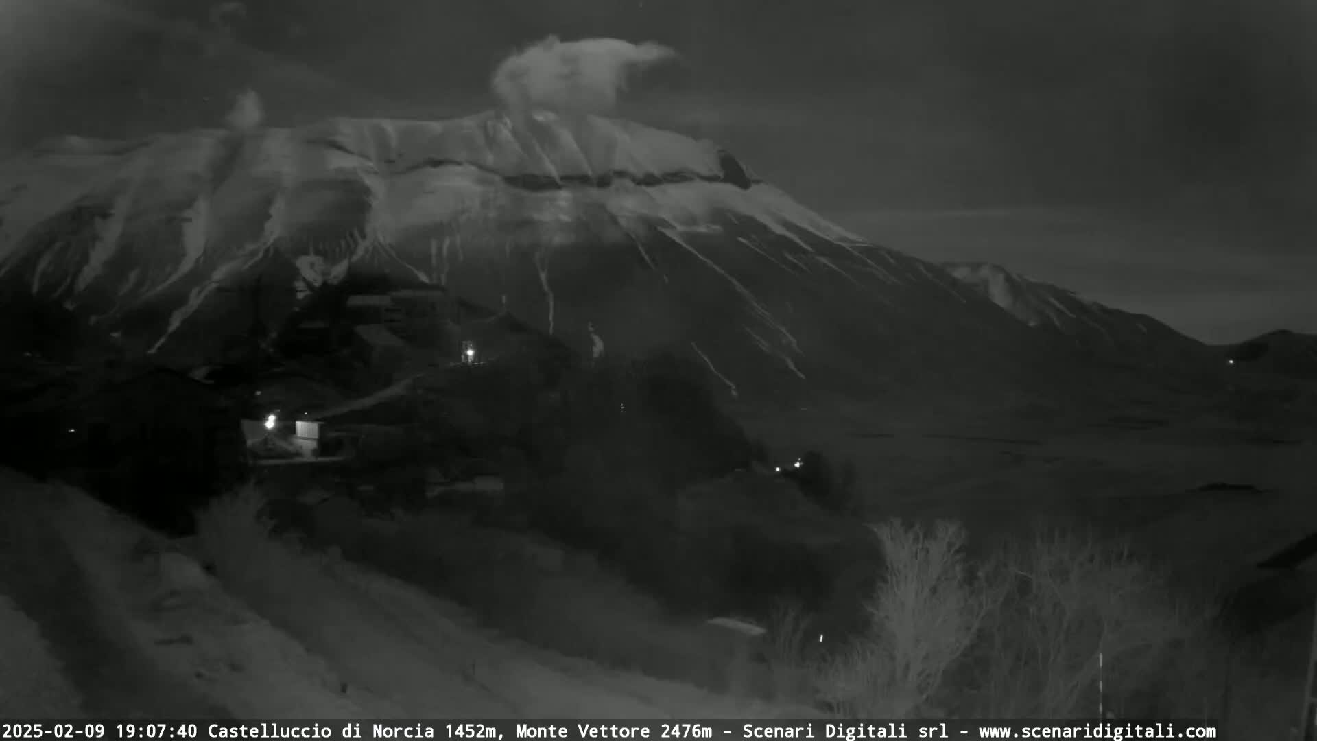 A snow-covered mountain is visible at night, with a few lights illuminating a village in the foreground.