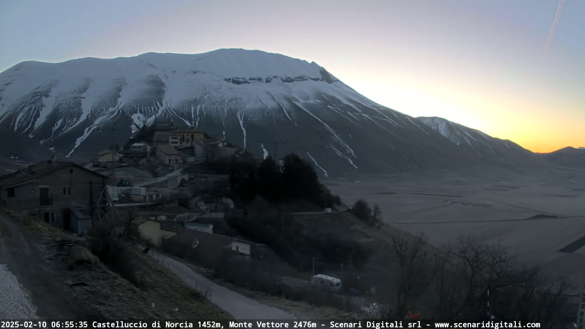 A snow-capped mountain looms over a small village at sunrise, with a valley stretching out in the foreground.