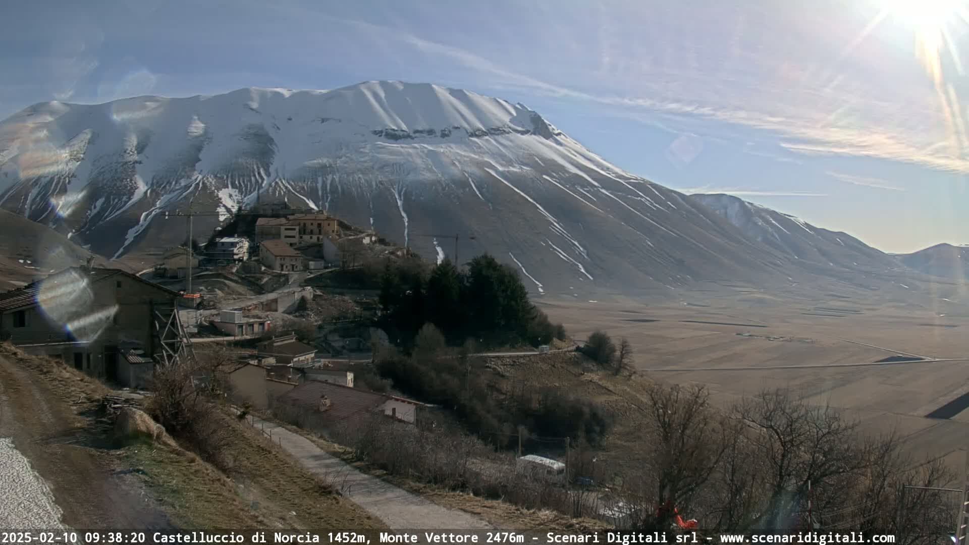 A snow-capped mountain overlooks a small village nestled in a valley on a sunny day.