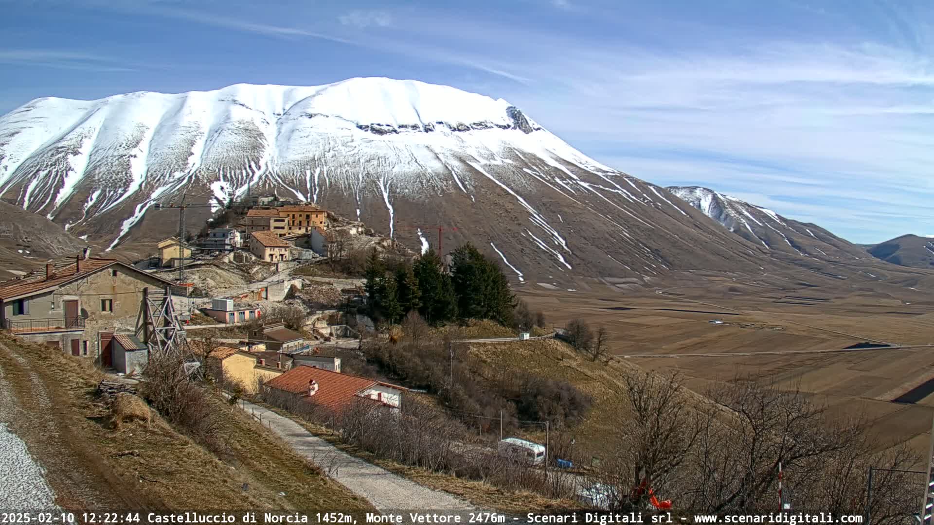 A snow-capped mountain overlooks a small village nestled in a valley of brown fields under a clear blue sky.