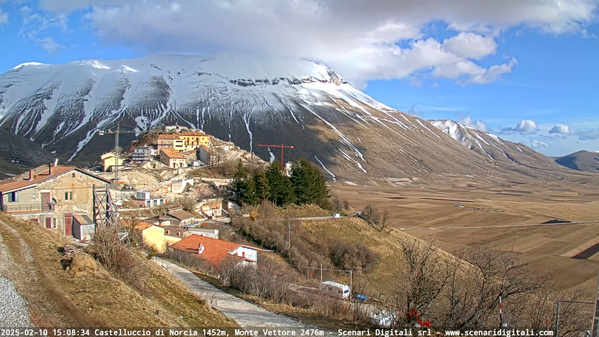 A partly sunny day reveals a snow-capped mountain overlooking a small village nestled in a valley, with construction underway and a brown, expansive plain stretching into the distance.