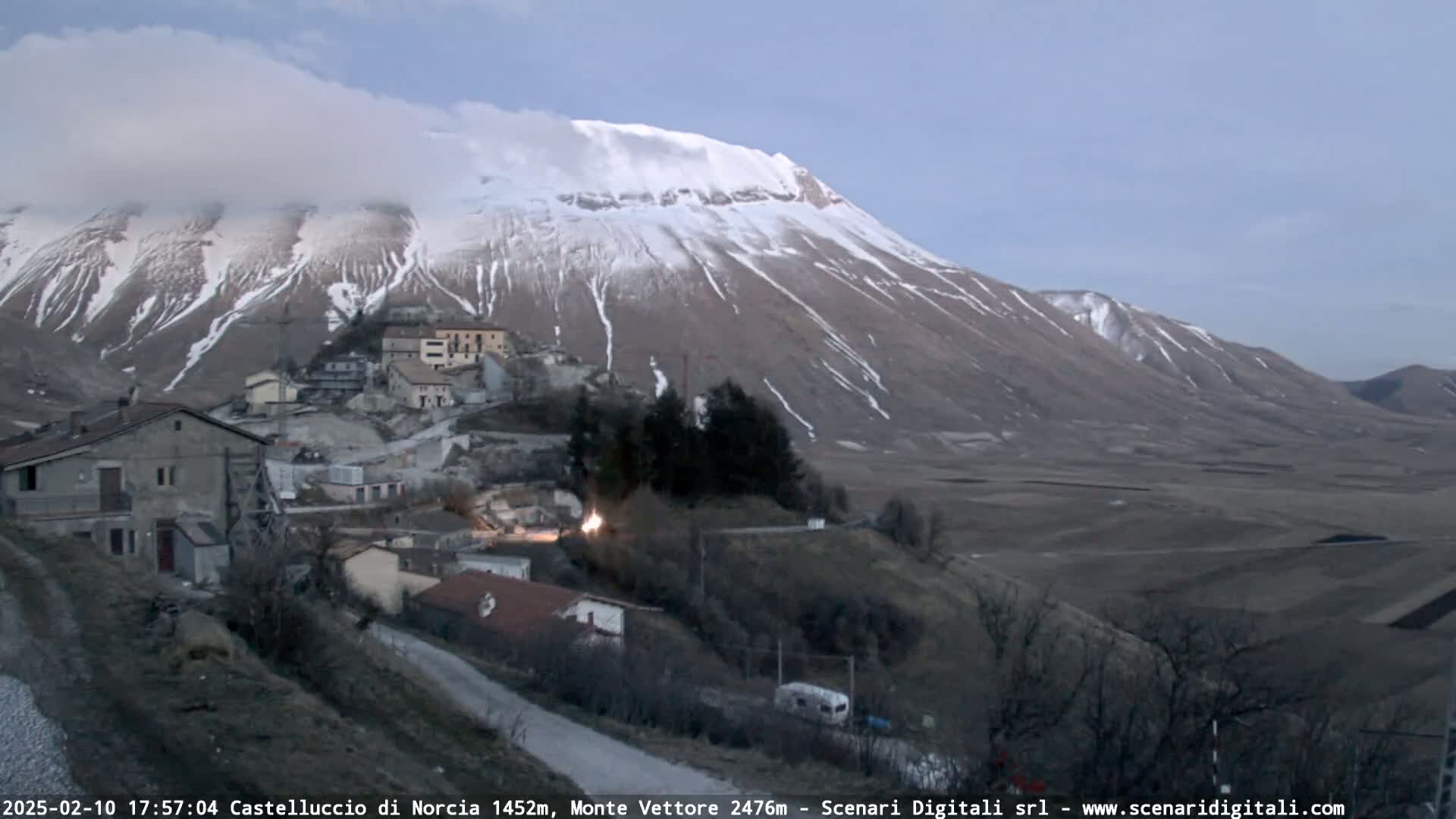 A snow-dusted mountain looms over a small village nestled in a valley, under a twilight sky.