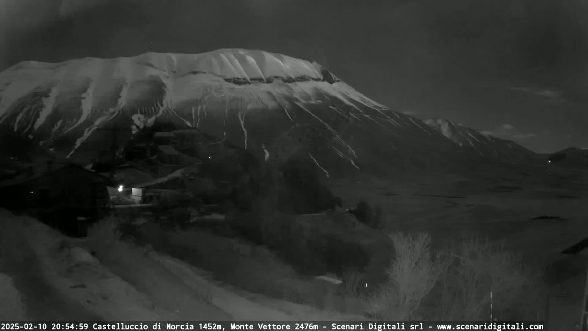 A snow-covered mountain looms over a small village at night under a dark sky.