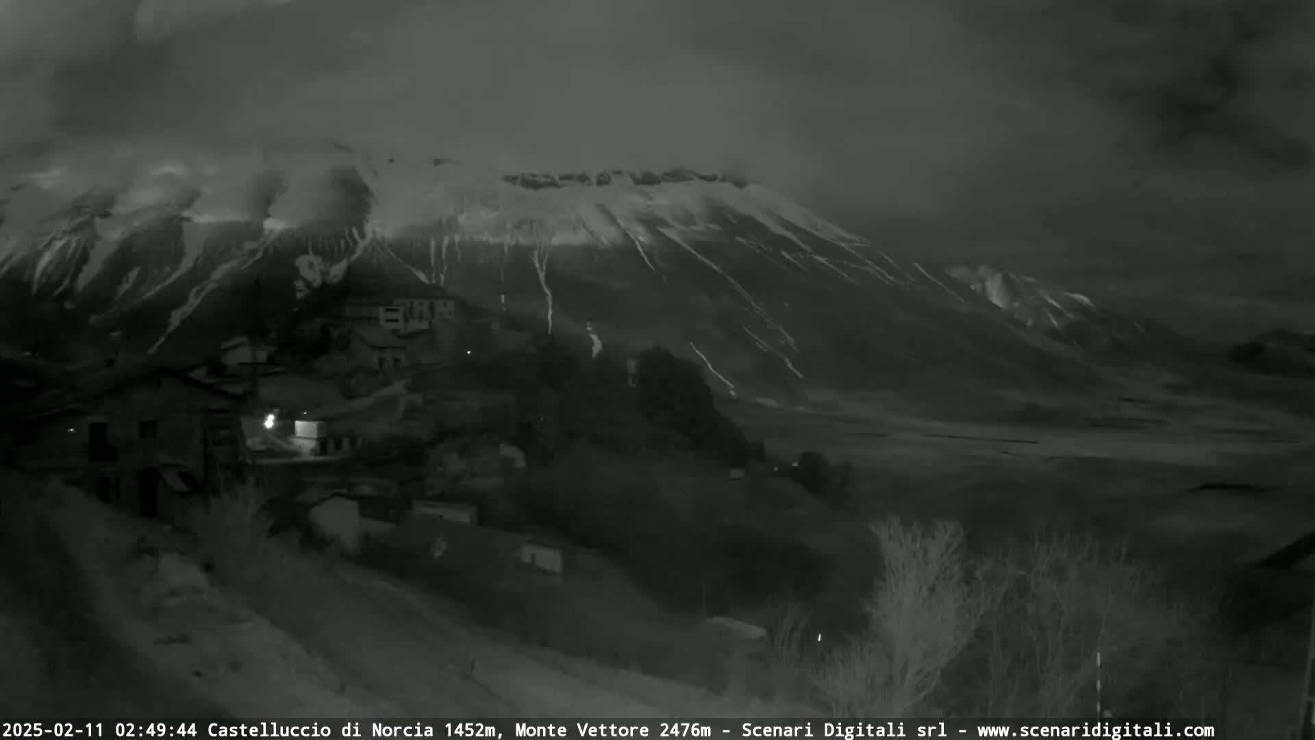 A nighttime, low-light view shows a snow-covered mountain overlooking a small village under a dark, cloudy sky.