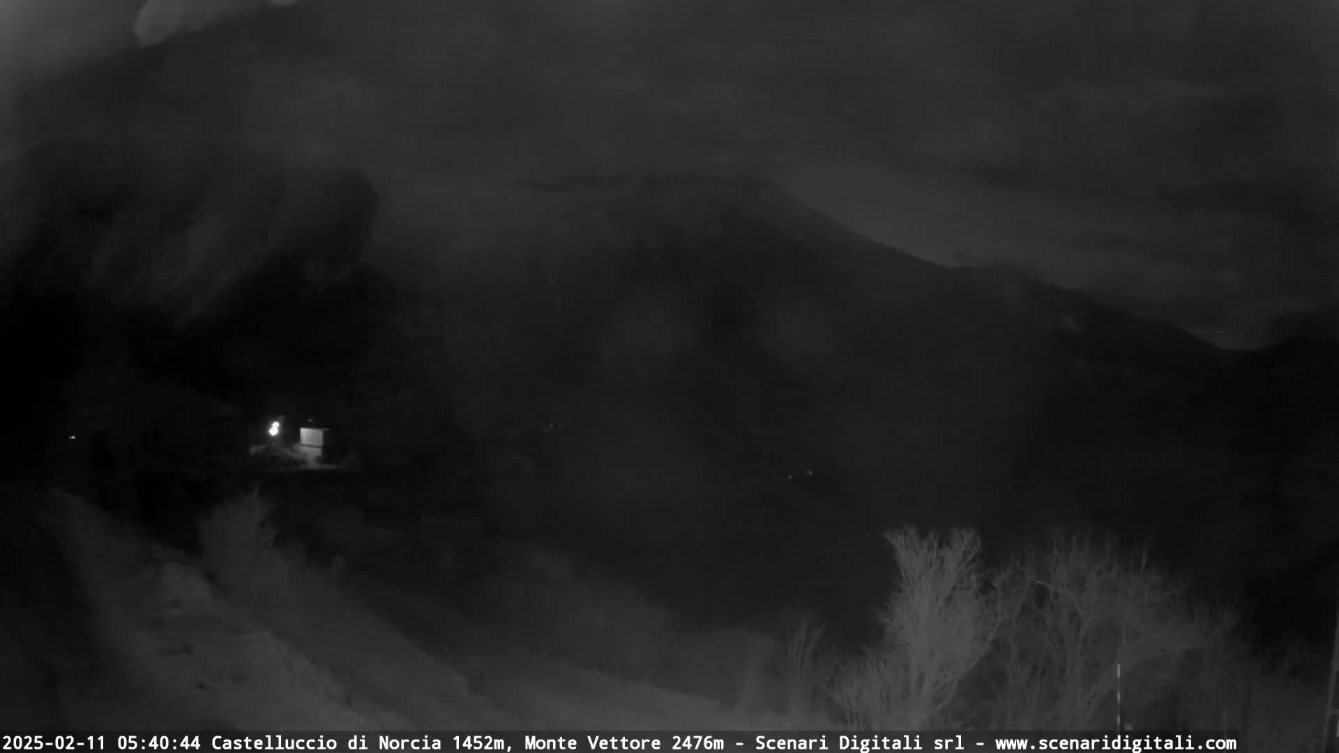 A nighttime, low-light view of a mountainous landscape under a cloudy sky, with a few small lights visible in the foreground.