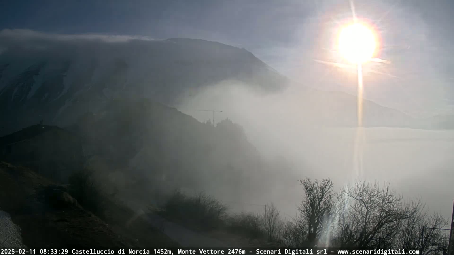 A bright sun shines through a thick fog covering a mountainous landscape with a construction crane visible in the middle ground.