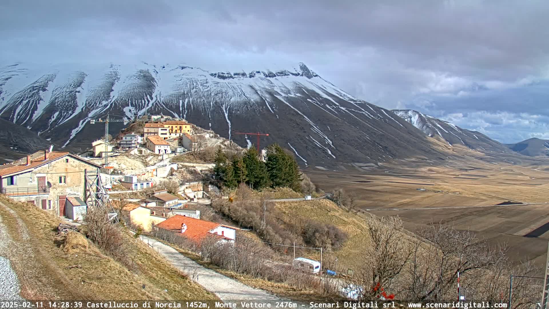 A small village nestled on a hillside, with construction cranes visible, overlooks a broad, flat valley and a snow-capped mountain range under a partly cloudy sky.