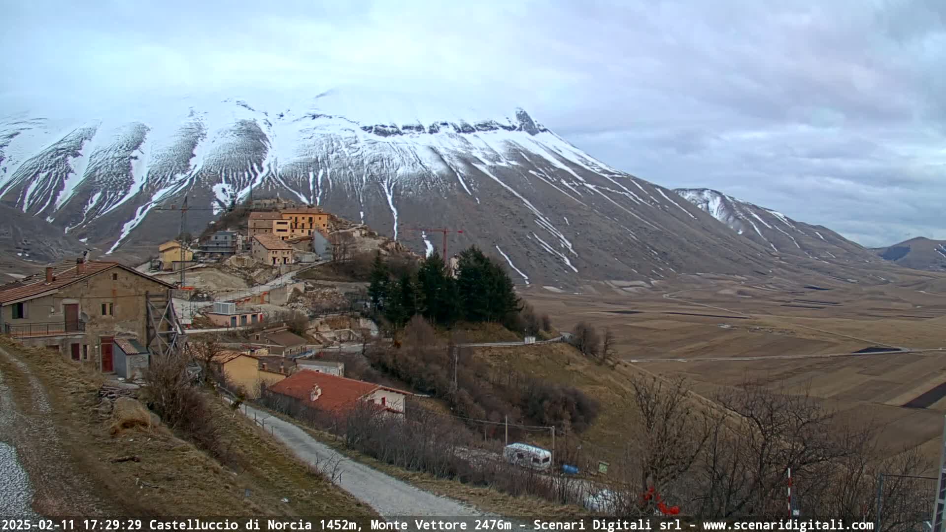 A snow-dusted mountain overlooks a small village nestled in a valley under a cloudy sky.