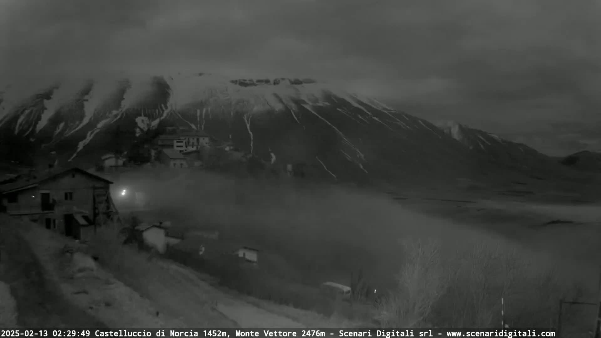A snow-dusted mountain looms over a foggy valley, with a small village nestled at its base under a dark, cloudy sky.