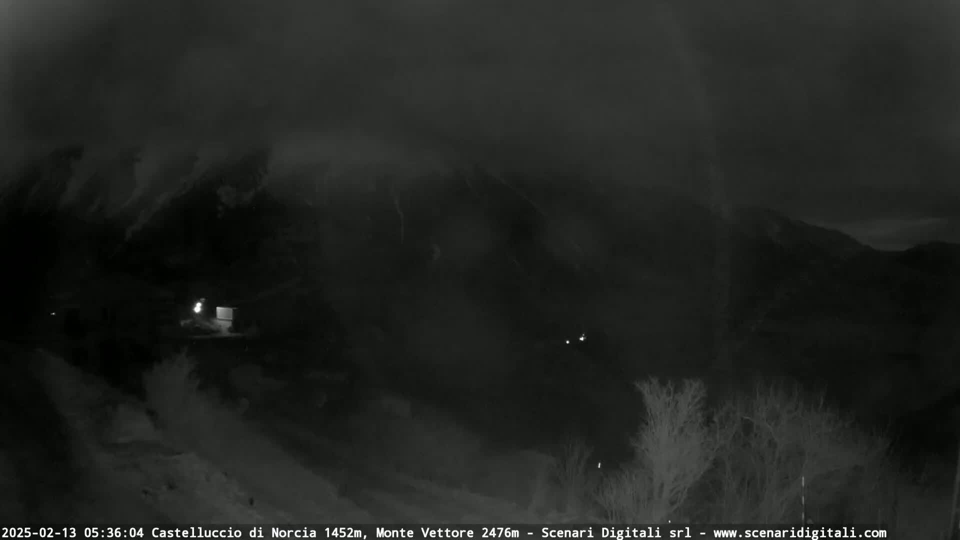 A nighttime, low-visibility view of a mountain range shrouded in fog or mist, with sparse vegetation in the foreground.