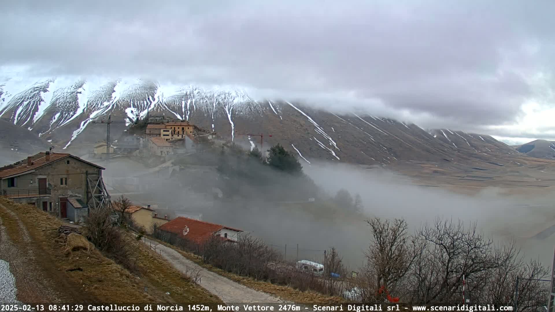 A foggy valley scene shows a small village nestled at the base of snow-dusted mountains under a cloudy sky.