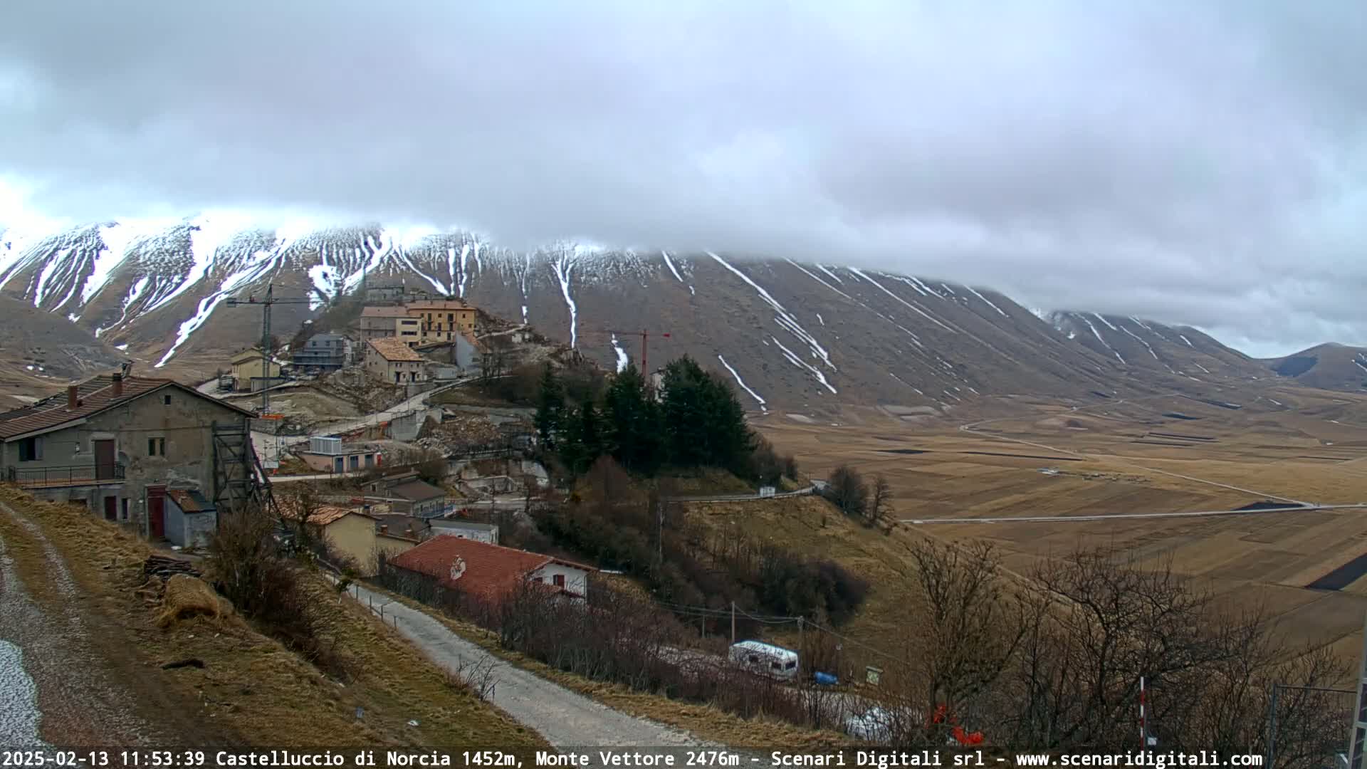 A small village nestled on a hillside overlooks a vast, flat plain under a cloudy sky, with snow-dusted mountains in the background.
