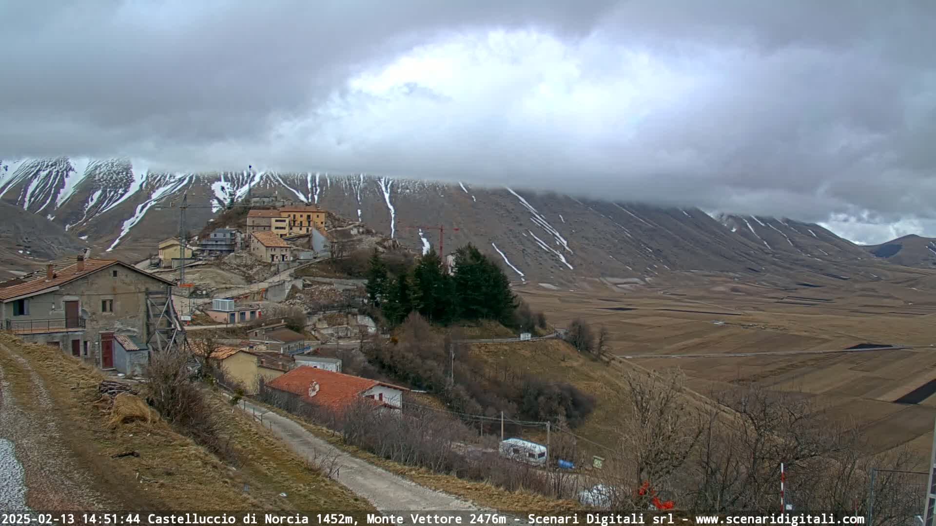 A small village nestled in a valley below snow-dusted mountains under a cloudy sky.