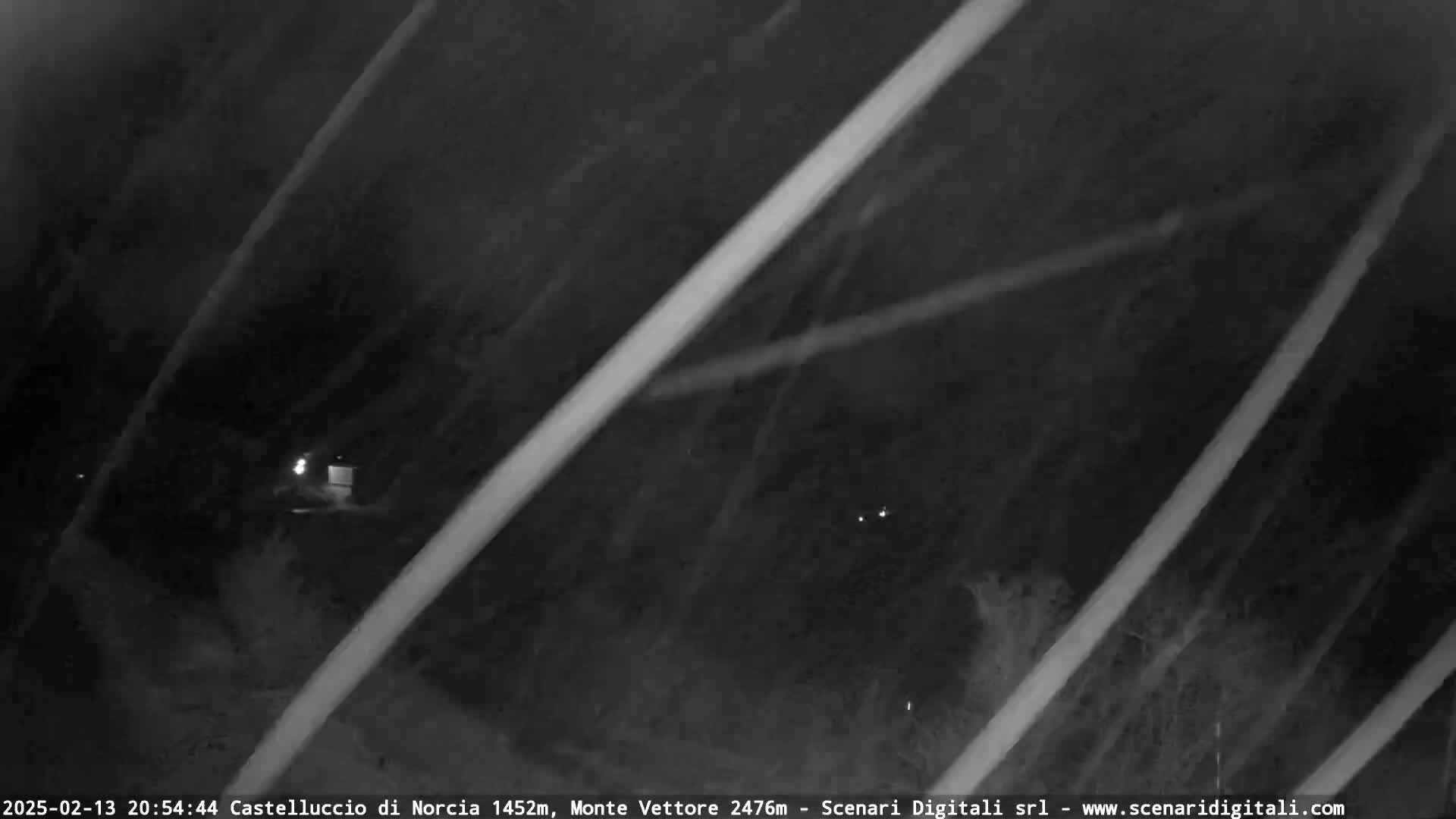 A nighttime, low-light image shows a dark, possibly snowy, landscape with thin, bright lines obscuring the view, and a few distant lights are visible.